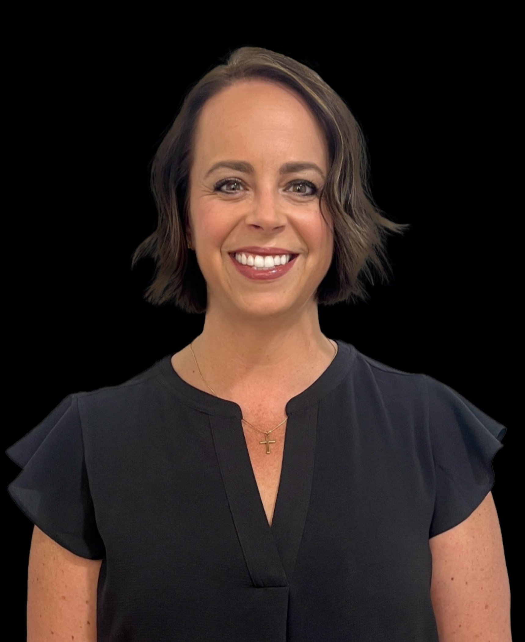A smiling woman with short brown hair, wearing a black top with ruffled sleeves, a gold cross necklace, and makeup, standing against a black background.