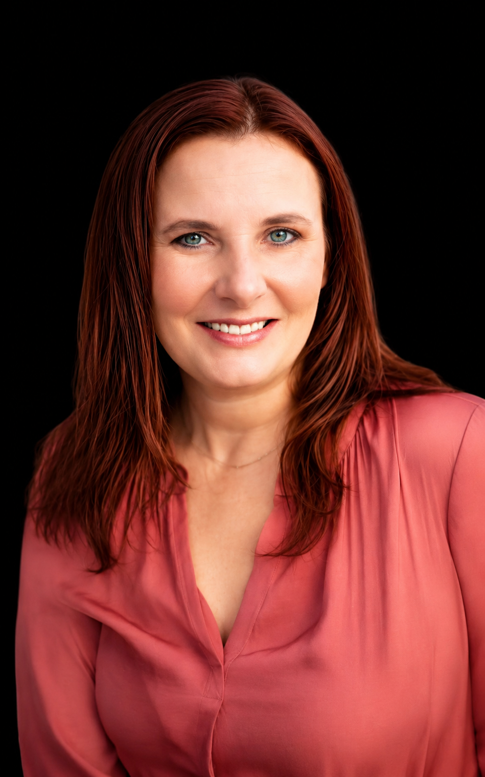 A woman with long red hair, blue eyes, and a light skin tone, wearing a pink blouse, smiling at the camera against a black background.