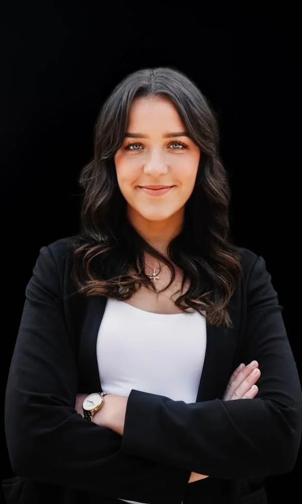 A smiling middle-aged woman with shoulder-length blonde hair and glasses, wearing a white blouse with a black cardigan, gold necklace, and earrings, standing against a black background.