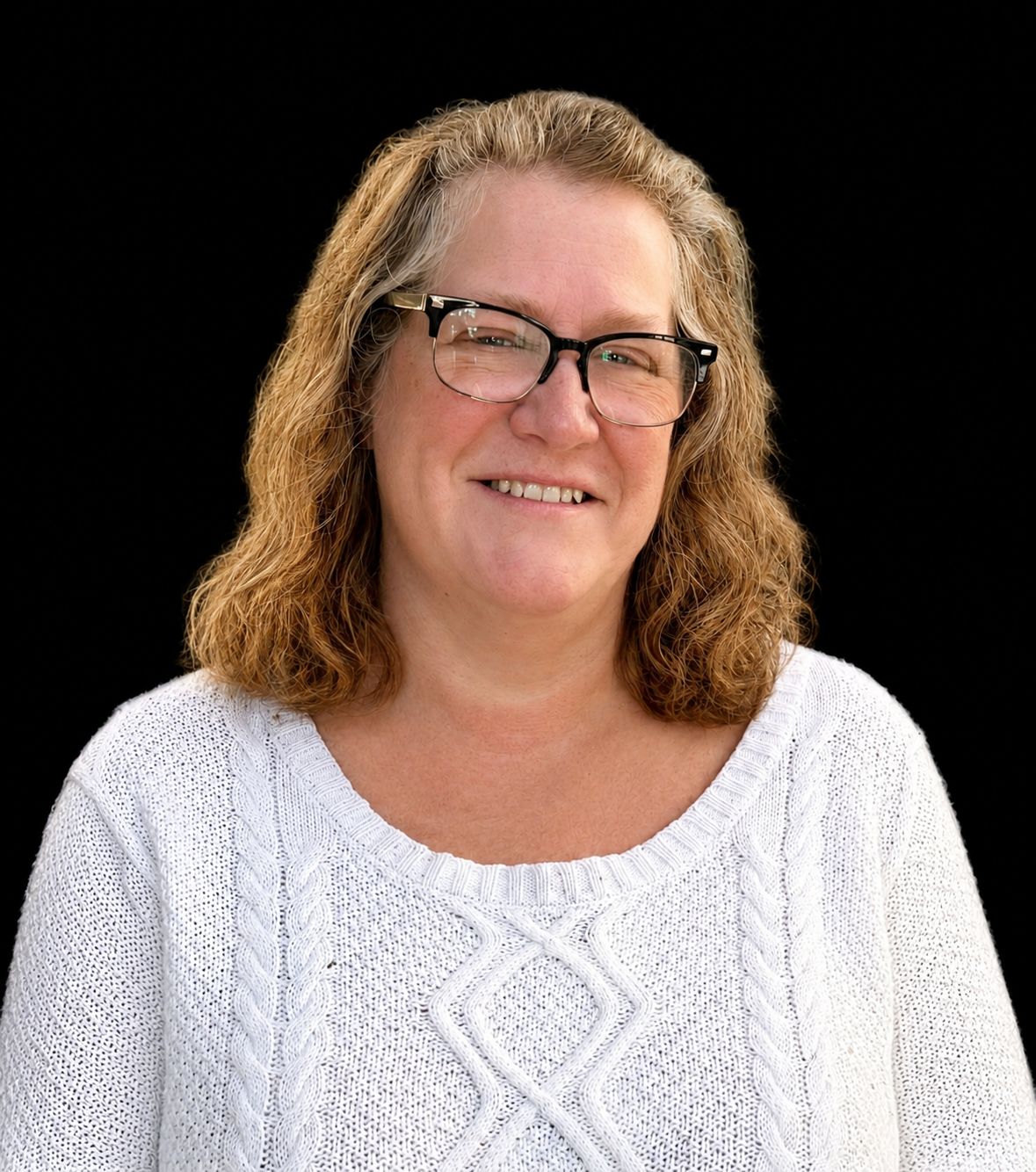 A woman with curly red hair, glasses, and a white sweater, smiling against a black background.