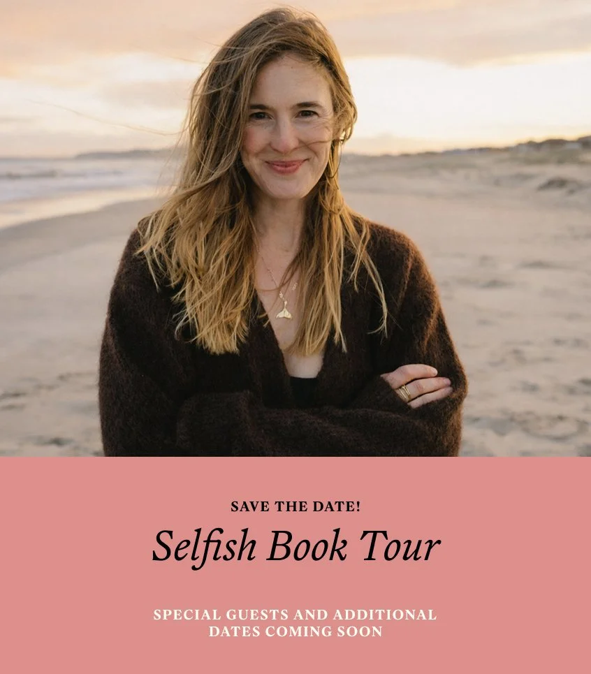 A woman with long, wavy, blonde hair smiling on a sandy beach at sunset, wearing a dark sweater and gold jewelry.