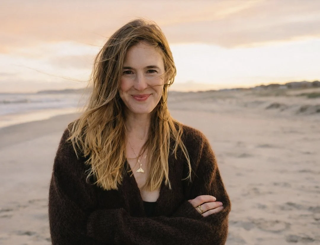 A woman with long, wavy brown hair smiling on a beach at sunset, wearing a dark sweater and layered necklaces.