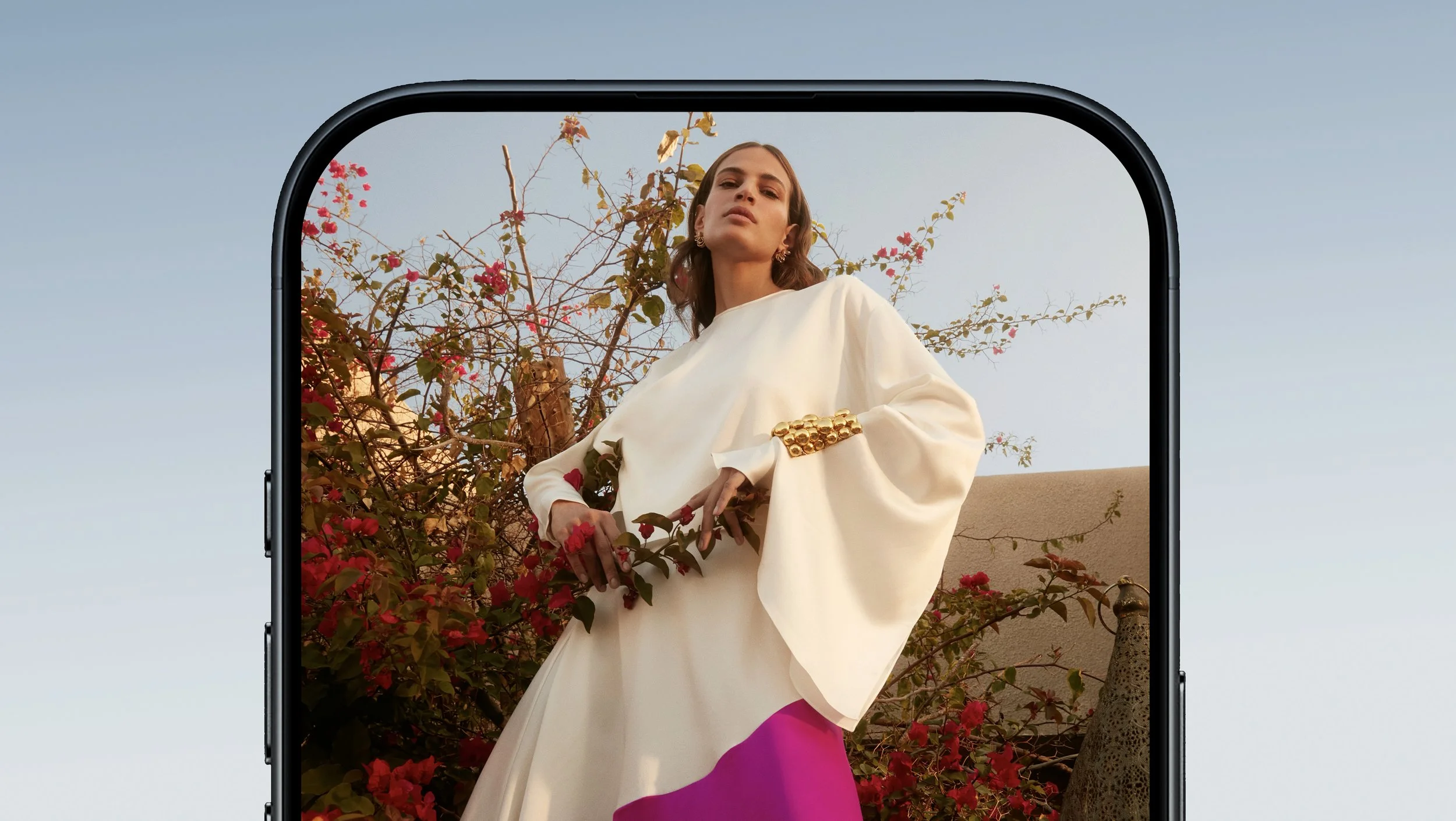 A woman in a flowing white dress with puffy sleeves and gold jewelry stands outdoors near flowering bushes with pink and red flowers, viewed through the screen of a smartphone.