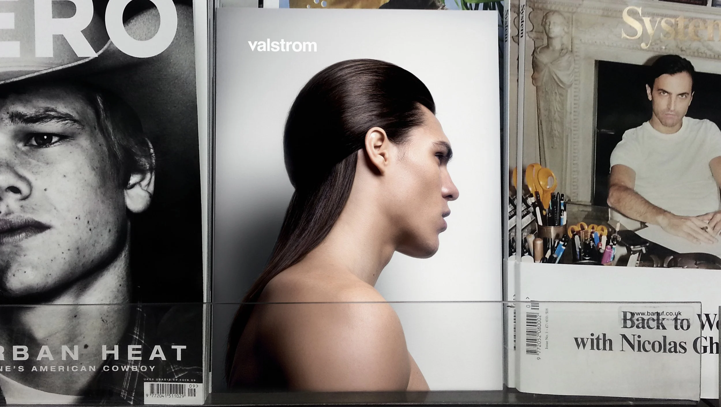 Side view of a young man with long, dark, slicked-back hair, shirtless, sitting at a desk with various stationery items, in front of a mirror with an ornate frame. On the left, part of a magazine cover with a black-and-white photo of a man's face and the text 'URBAN HEAT'. On the right, part of another magazine cover featuring a man in a white shirt in front of a mirror.