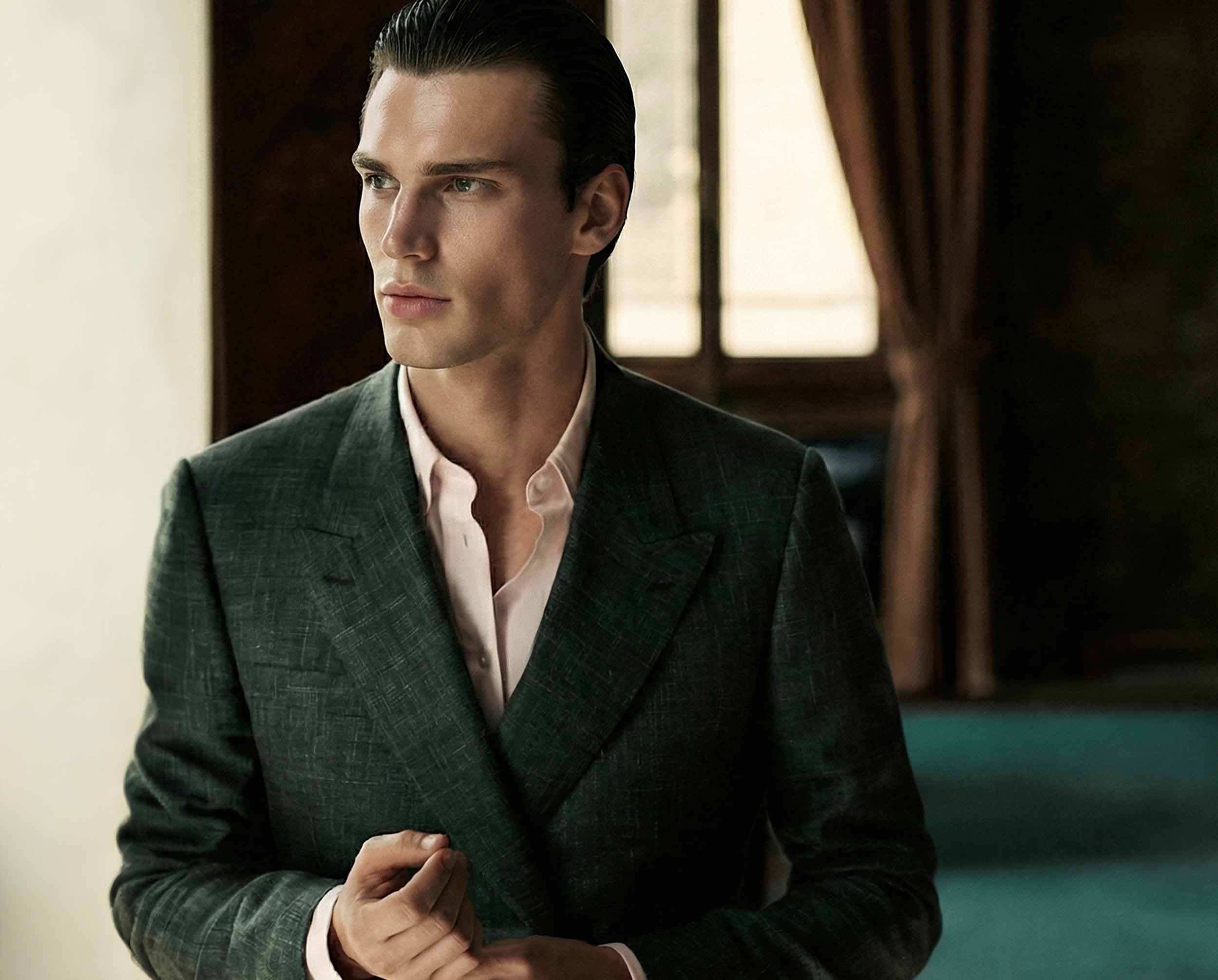 A young man with slicked-back dark hair, dressed in a dark blazer and light pink shirt, sitting indoors near a window with brown curtains in the background.