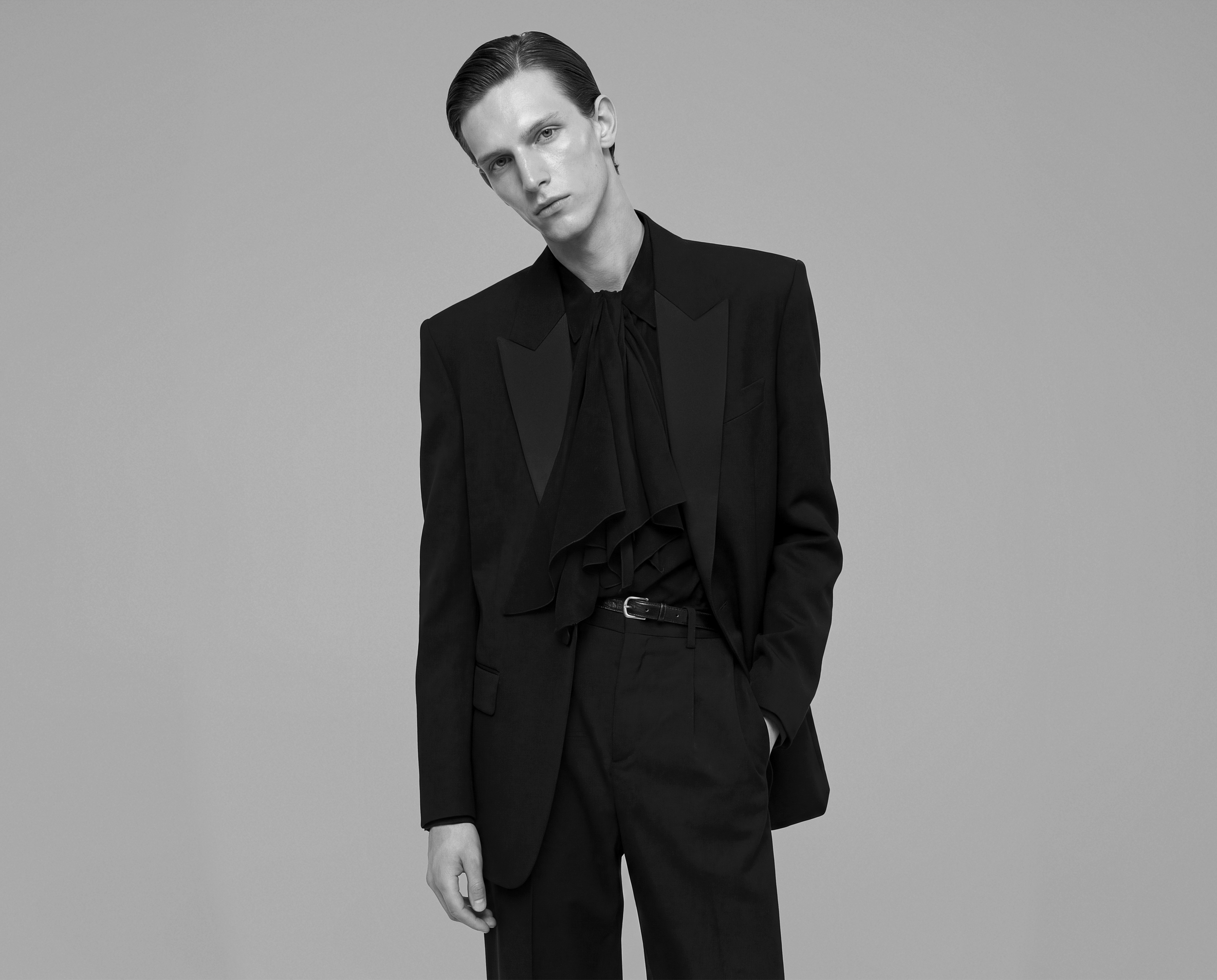 Black and white portrait of a young man with slicked-back hair wearing a tailored black suit with a ruffled black shirt, standing against a plain background.