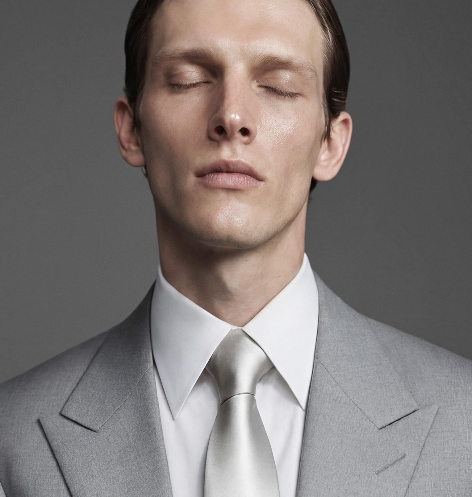 A young man with slicked-back hair in a closed-eye pose, wearing a gray suit, white shirt, and matching gray tie, against a gray background.