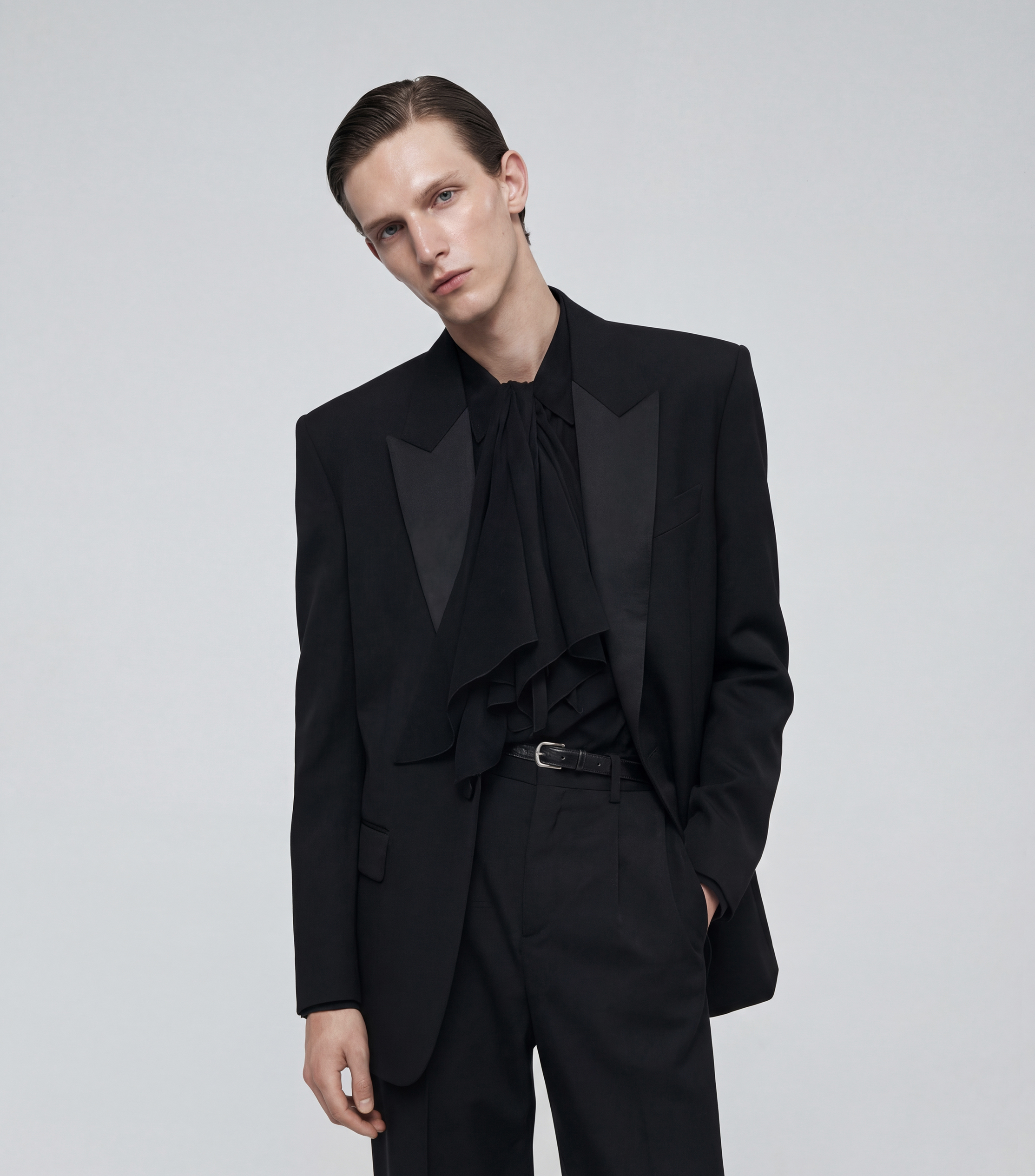 A young man with slicked back hair wearing a black suit with a ruffled shirt underneath, standing against a plain light gray background.