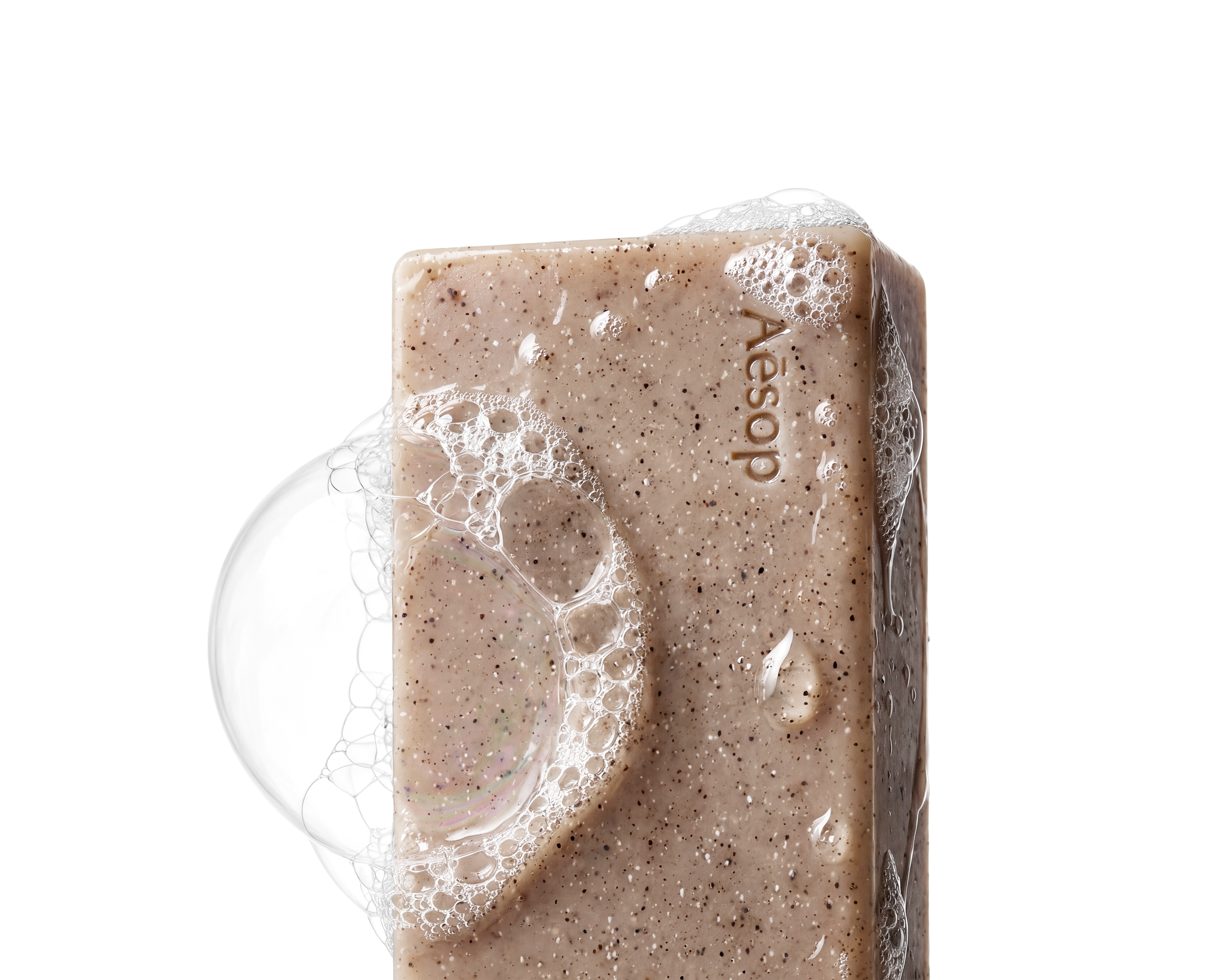 A close-up of a bar of beige soap with small dark specks, covered in soap bubbles, with the word 'Aesop' engraved on it.