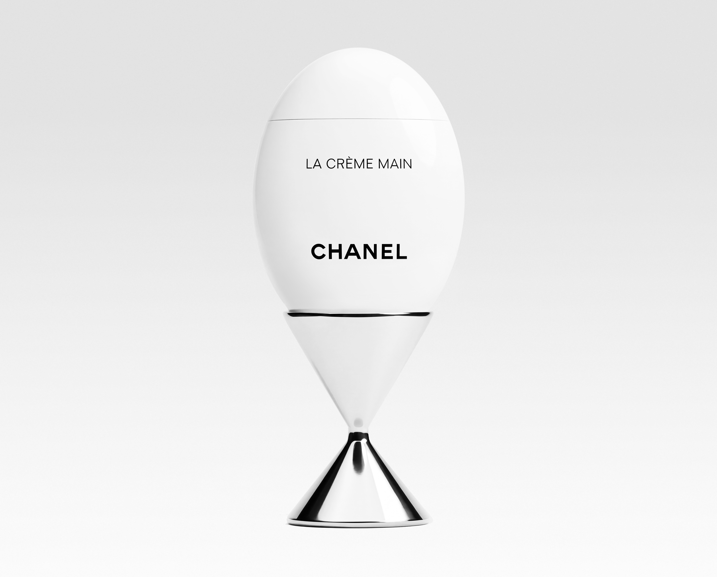 A white bottle labeled 'LA CRÈME MAIN' and 'CHANEL' with a black cap and a thin black line around the middle, reflected on a glossy surface.