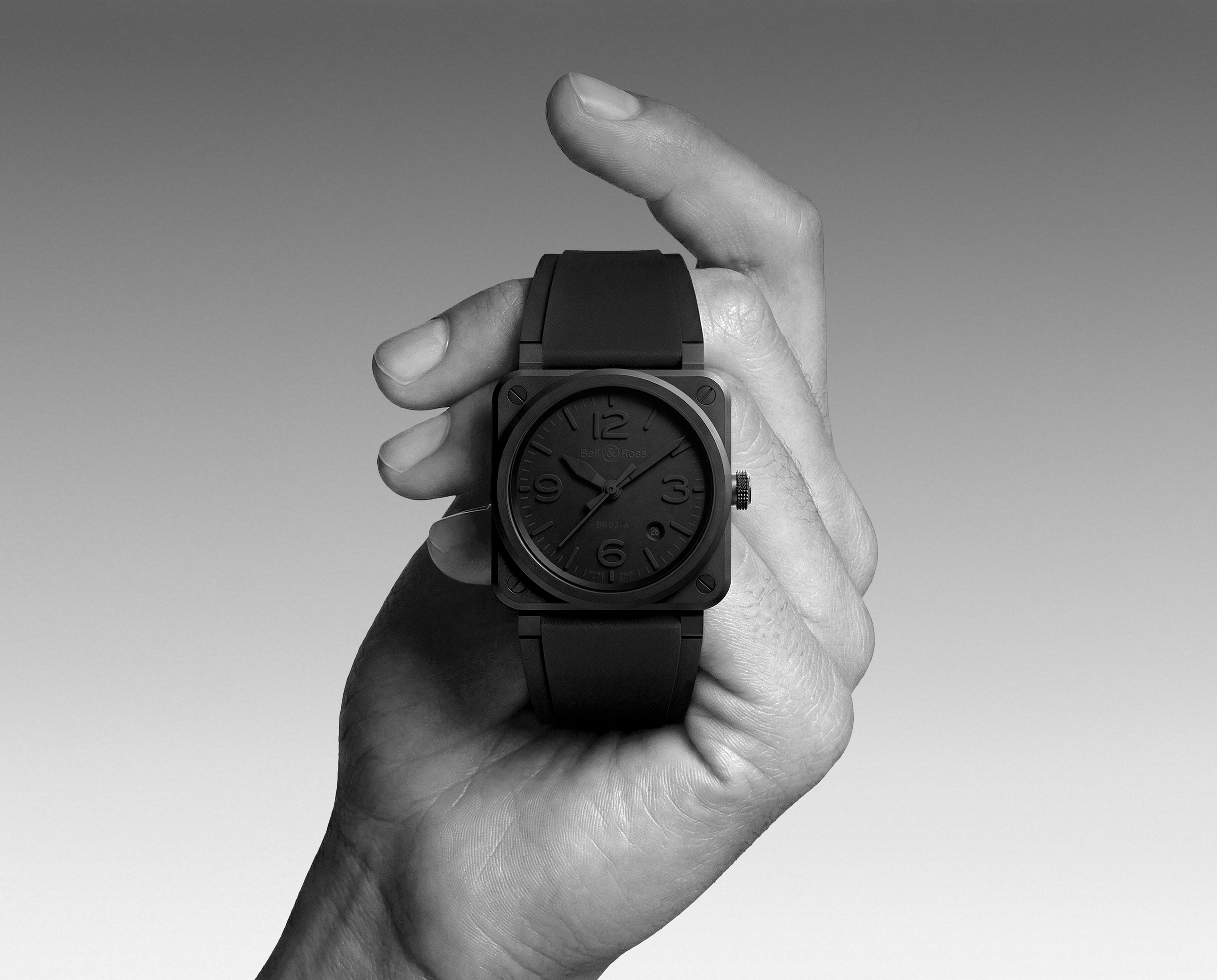 A person's hand holding a large, black wristwatch with a square face against a plain background.