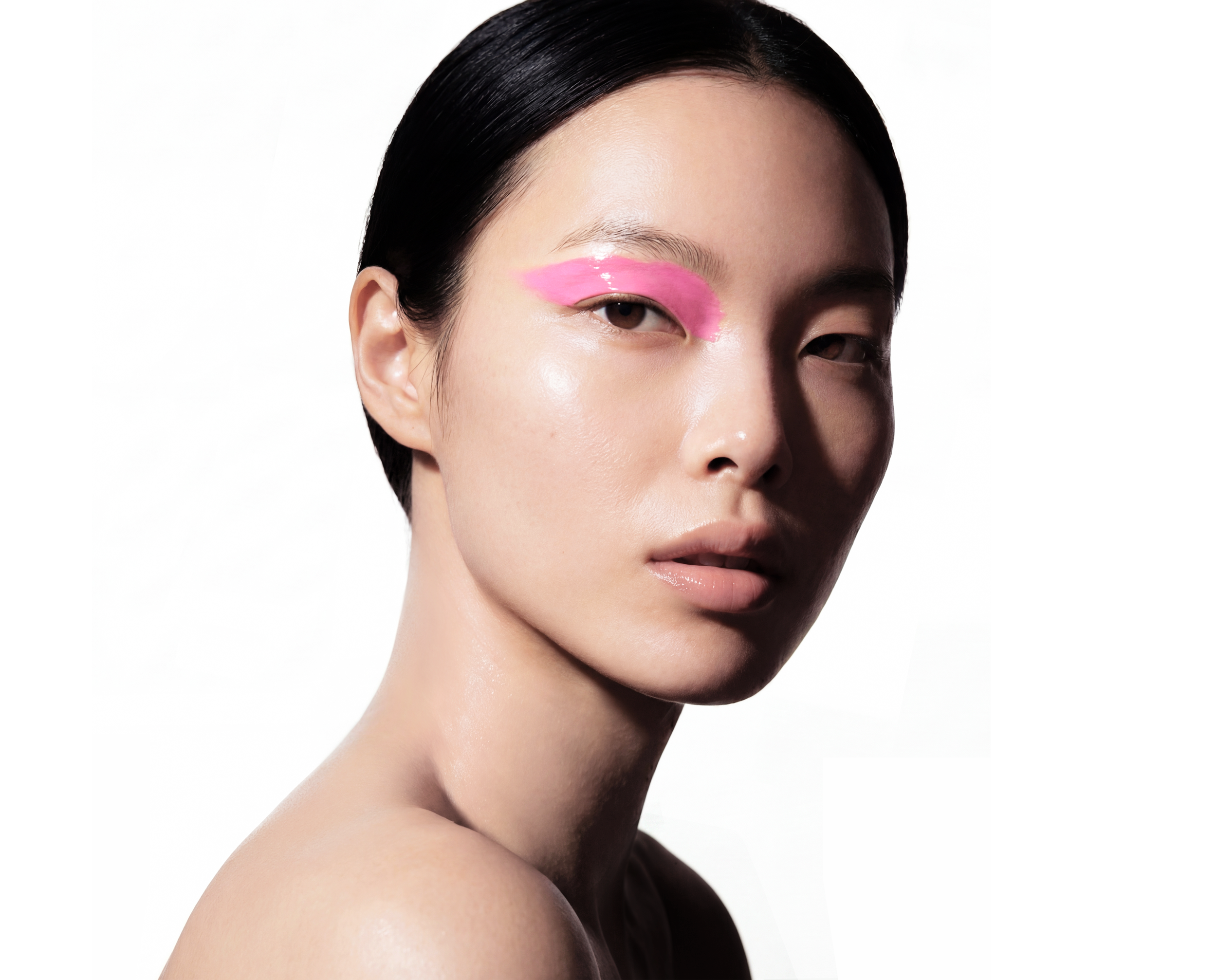 A close-up portrait of a woman with dark hair slicked back, showing her face and neck against a white background. She has pink eye makeup on her eyelids.