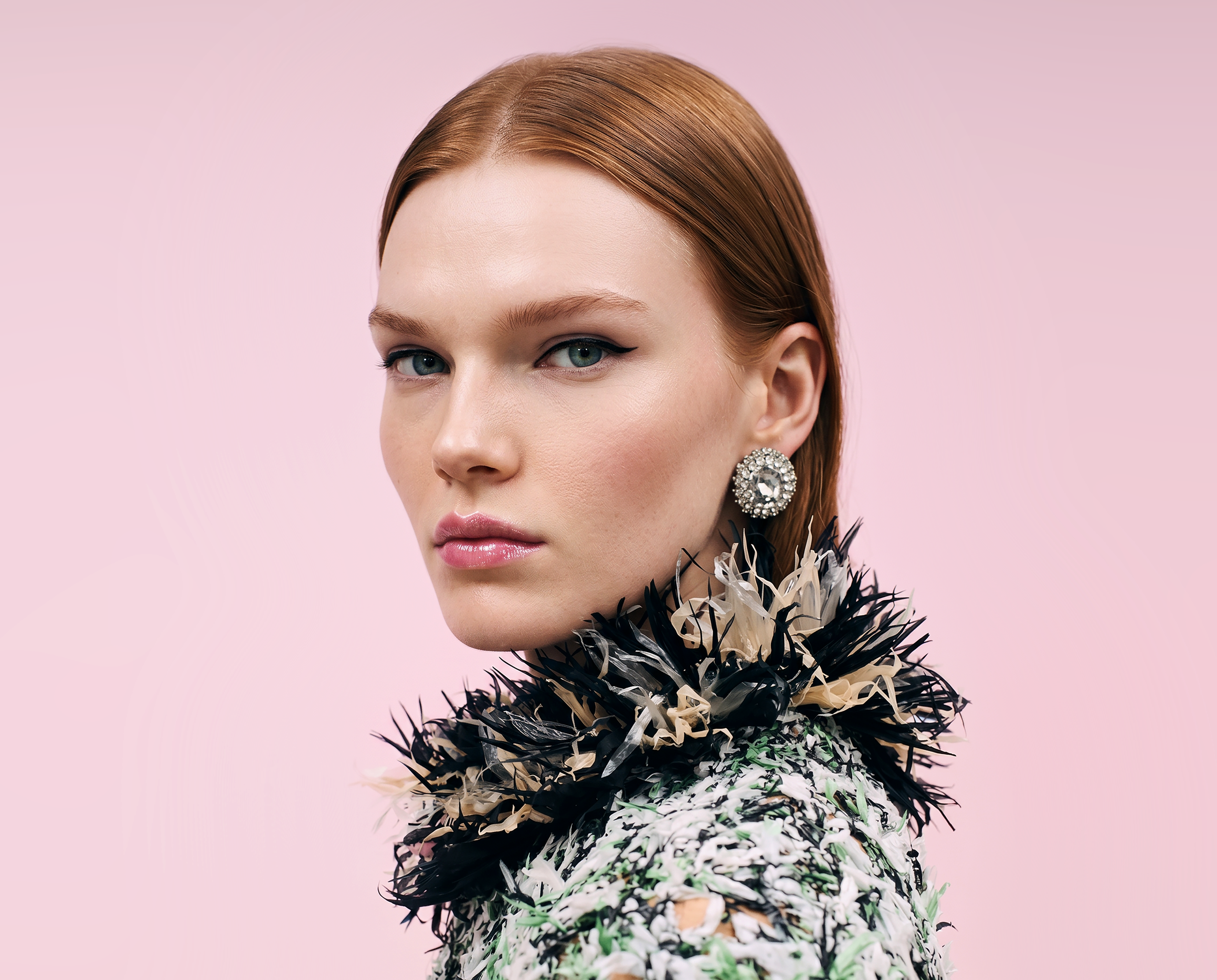 Close-up of a woman with red hair, wearing large jeweled earrings, a black and white feathered top, and makeup with winged eyeliner, against a pink background.