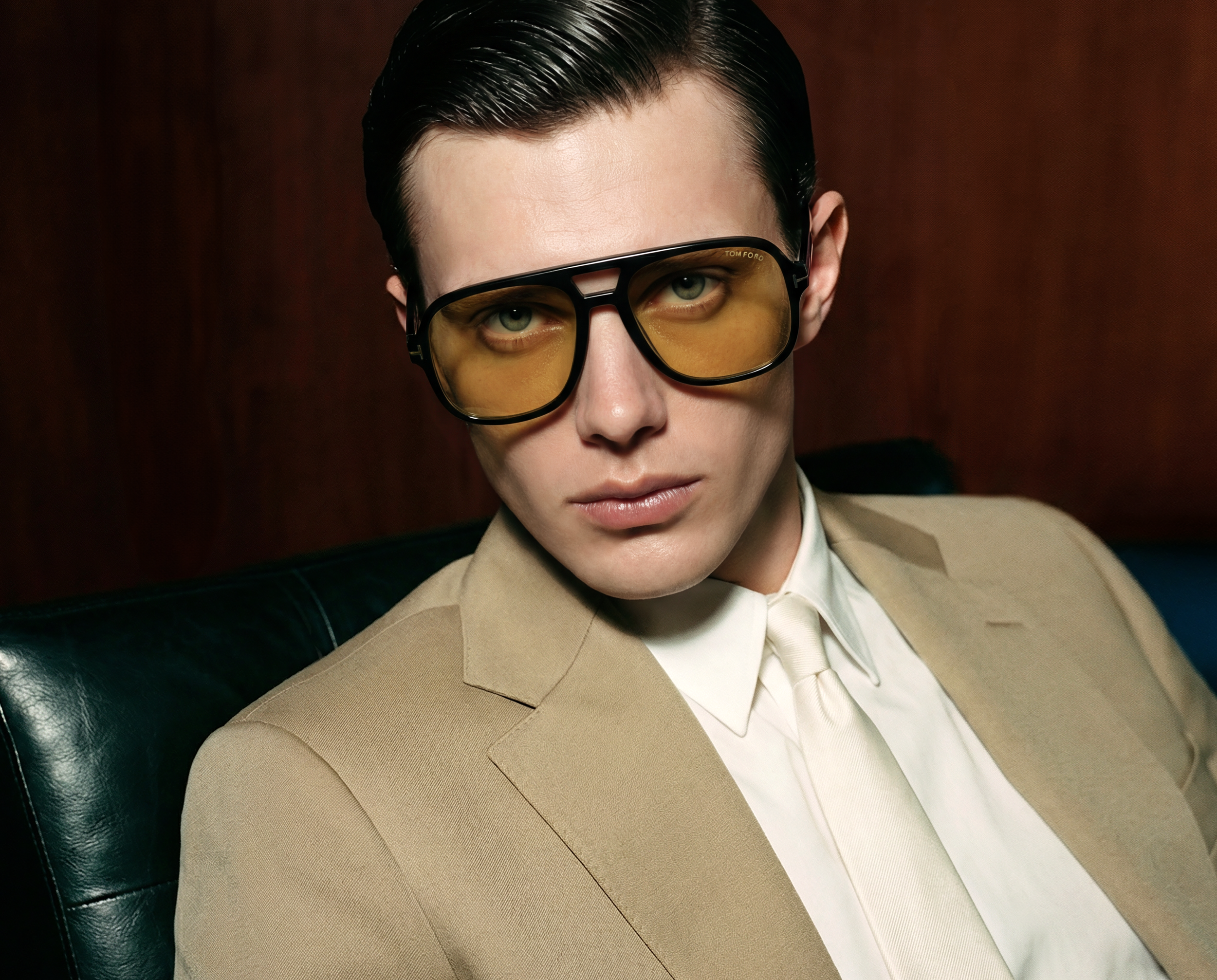Close-up of a man with slicked-back dark hair wearing large yellow-tinted Tom Ford glasses, tan blazer, and cream-colored shirt, sitting on a dark green leather chair with a brown background.