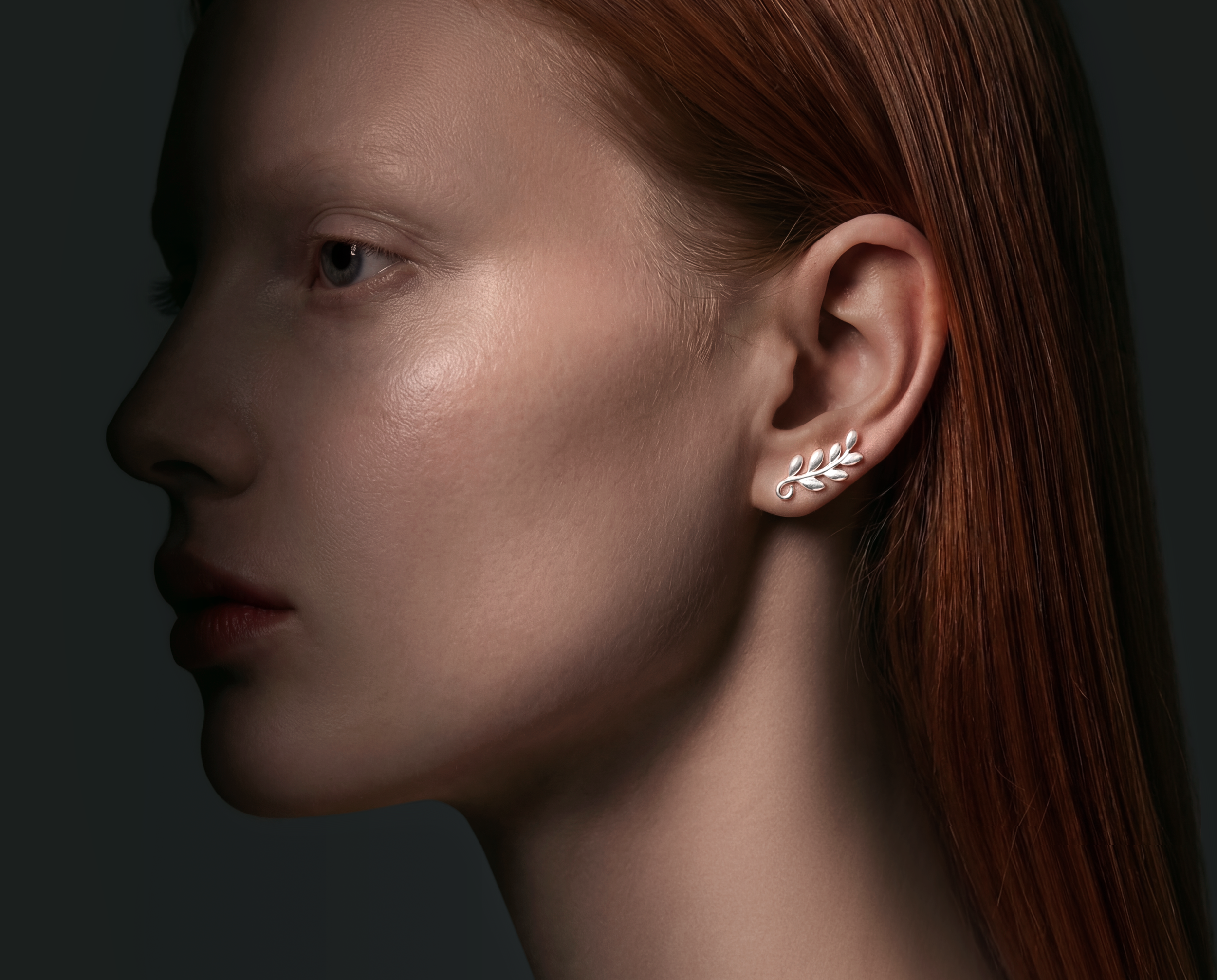 Close-up of a woman's face highlighting her skin, ear, and red hair with a silver leaf-shaped earring.