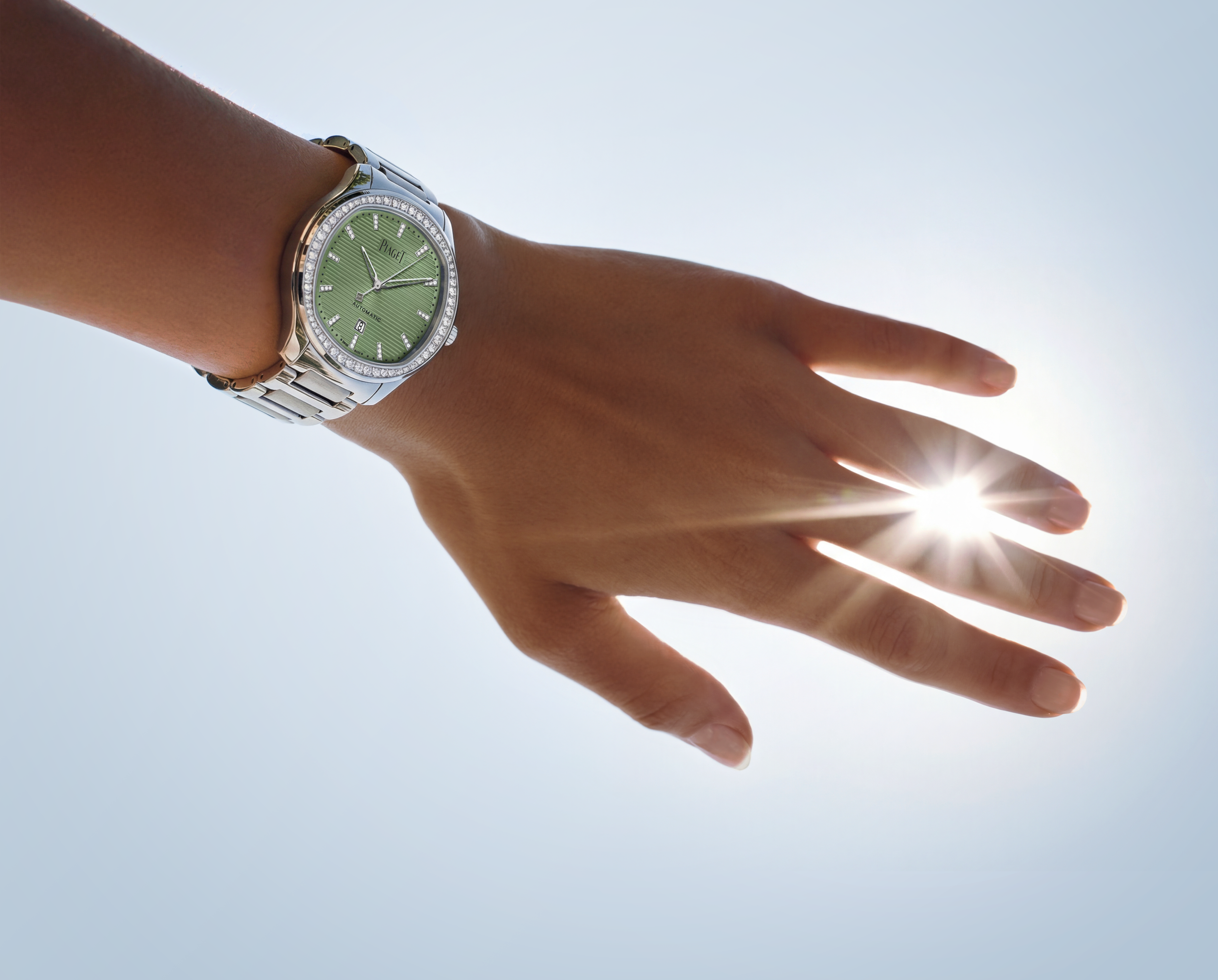 Close-up of a wristwatch with a green face and diamond accents on a person's wrist, held in front of a bright sky with sun rays.