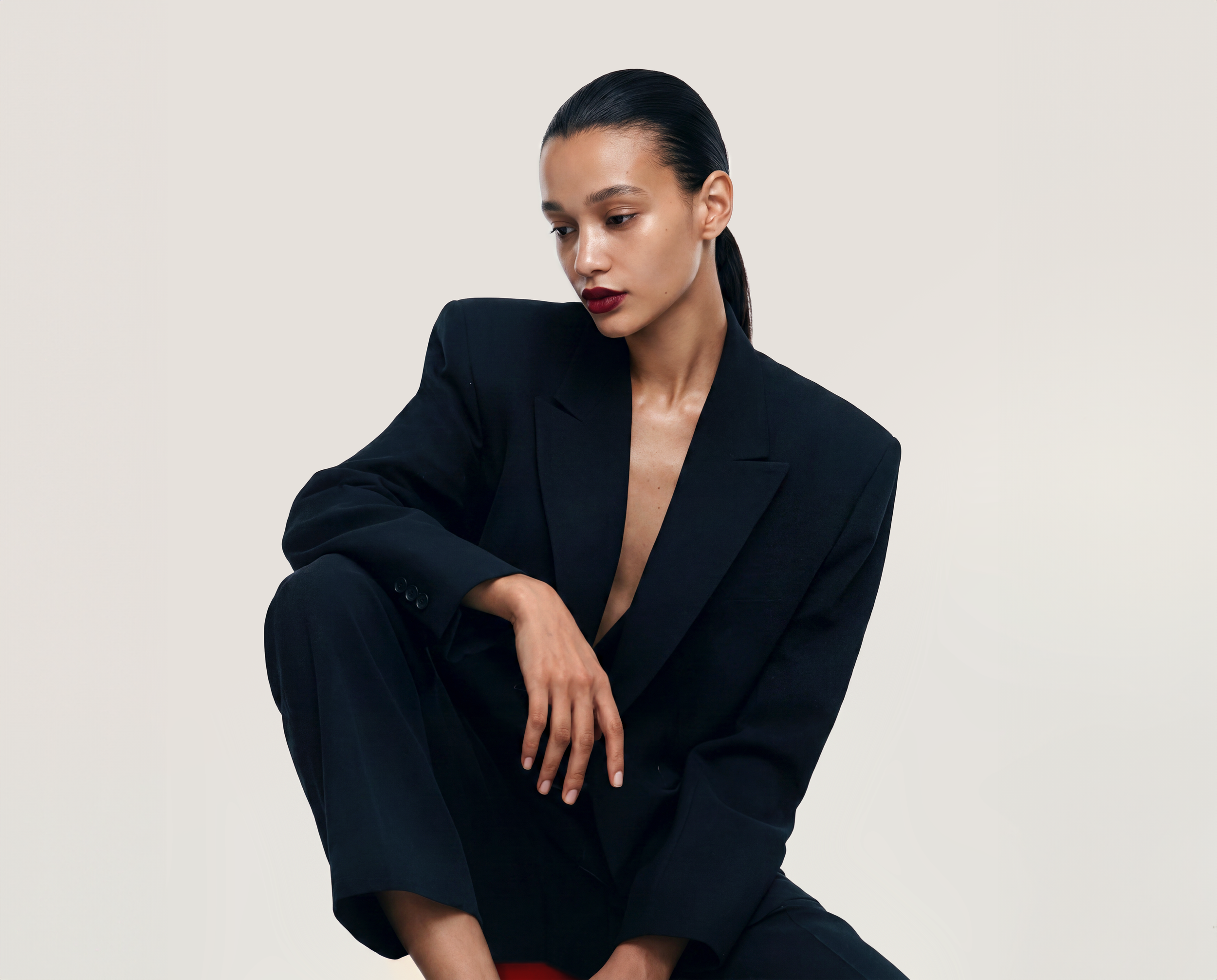 A woman with dark hair pulled back, wearing a black blazer and matching pants, sitting with her head slightly bowed and looking downward against a plain light background.