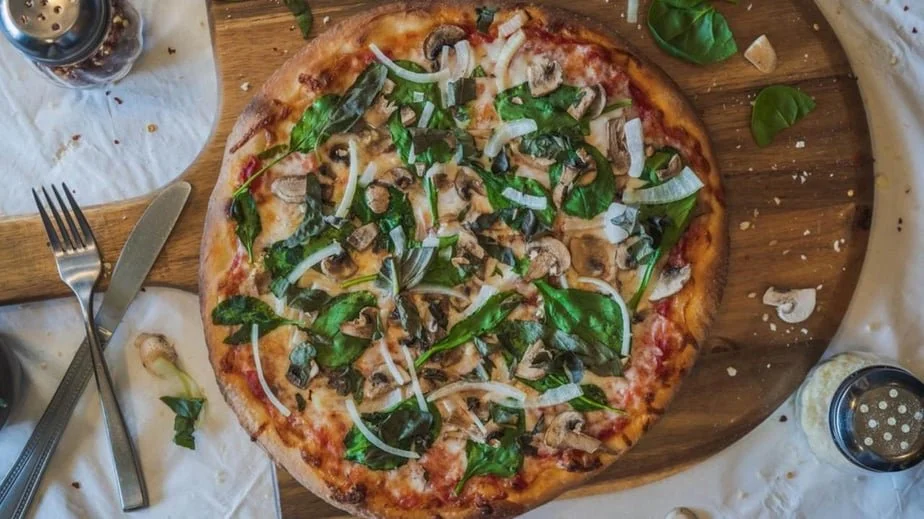 A mushroom and spinach pizza on a wooden serving board, with a metal pizza cutter nearby on a white tablecloth.