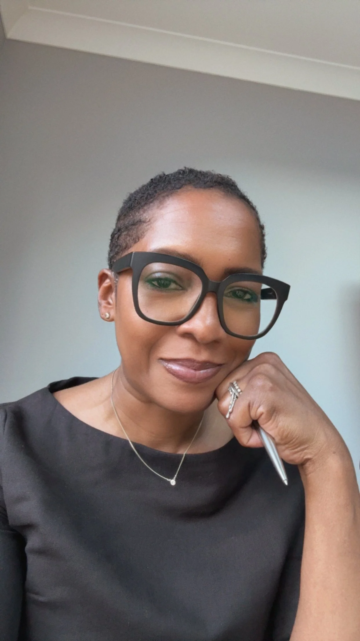 Chan Line of Fineline Counselling in Chalfont St Peter. A woman with short hair wearing large black glasses, a black top, a delicate necklace with a small pendant, earring studs, and a ring, smiling and resting her chin on her hand with a silver pen.