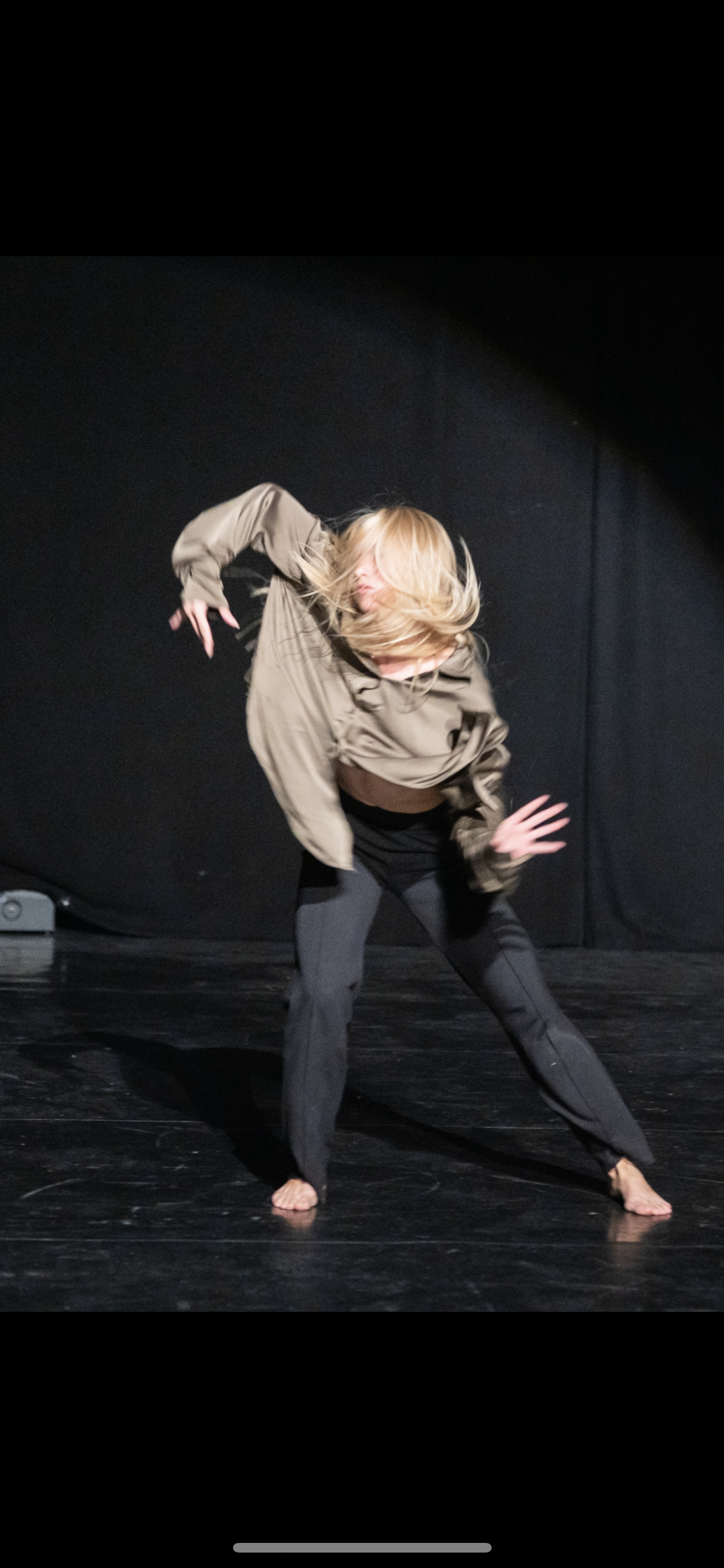 A woman with blonde hair dancing barefoot on stage, dressed in a loose brown jacket and black pants, with a black background.