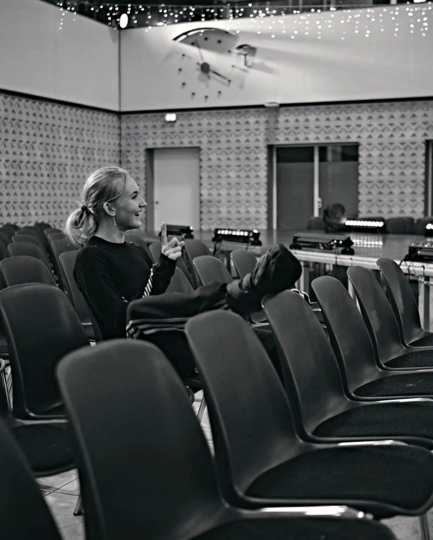 A woman sitting alone in an auditorium smiling and making a gesture with her hand. The auditorium has rows of empty chairs, a large clock on the ceiling, and stage lighting in the background.