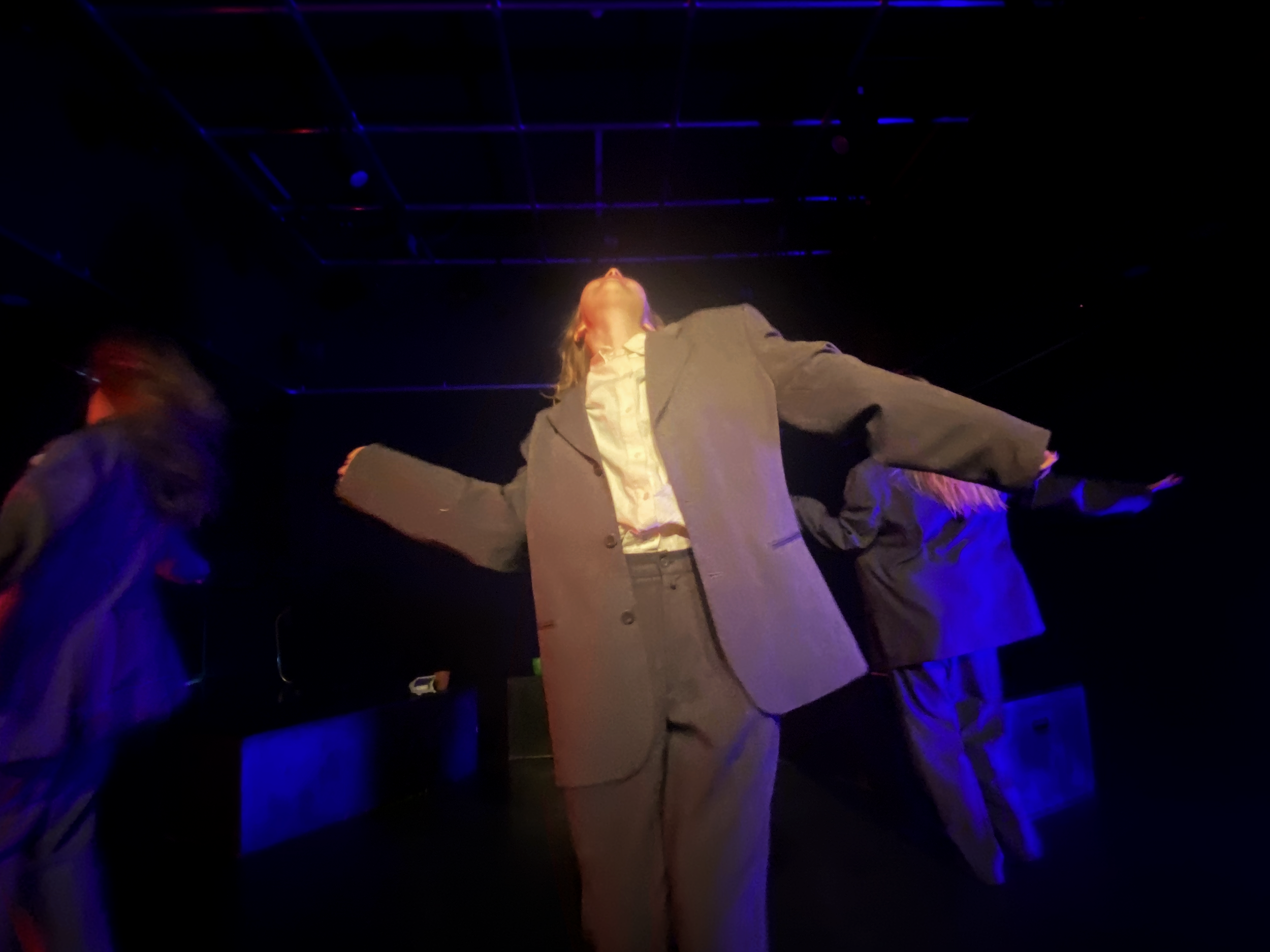 Person in a gray suit with arms outstretched on a stage with dark lighting and blue accents.