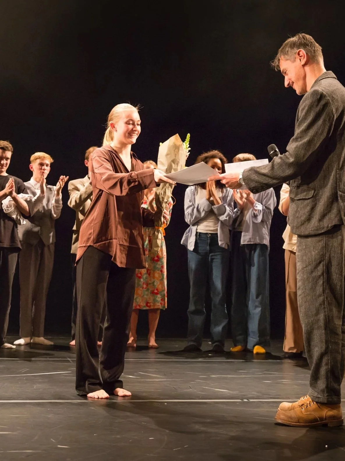 A young woman receiving an award and a bouquet of flowers on stage from a man in a suit, with several people standing and applauding in the background.