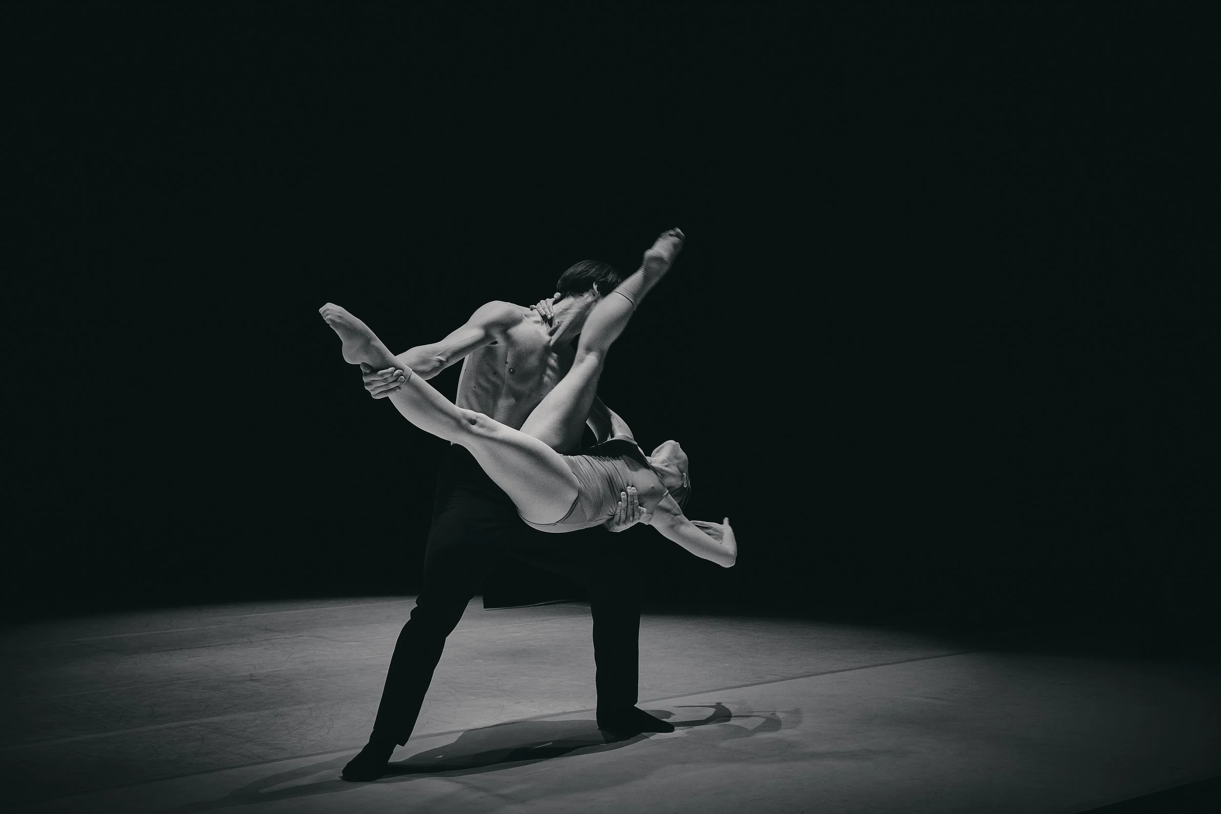 A male and female ballet dancers perform an acrobatic lift on stage, with a dark background.