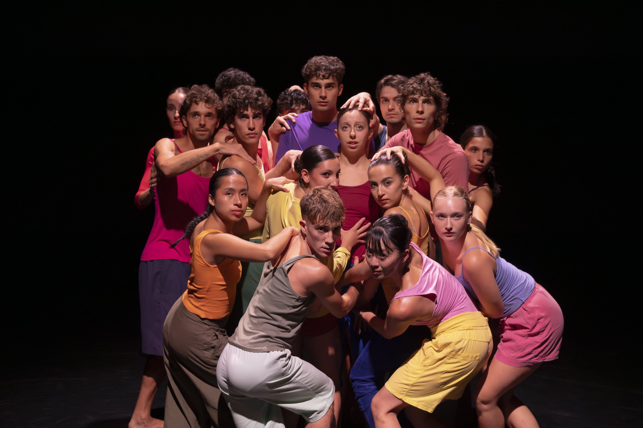 A group of young dancers in colorful casual clothing posing together on a dark stage.