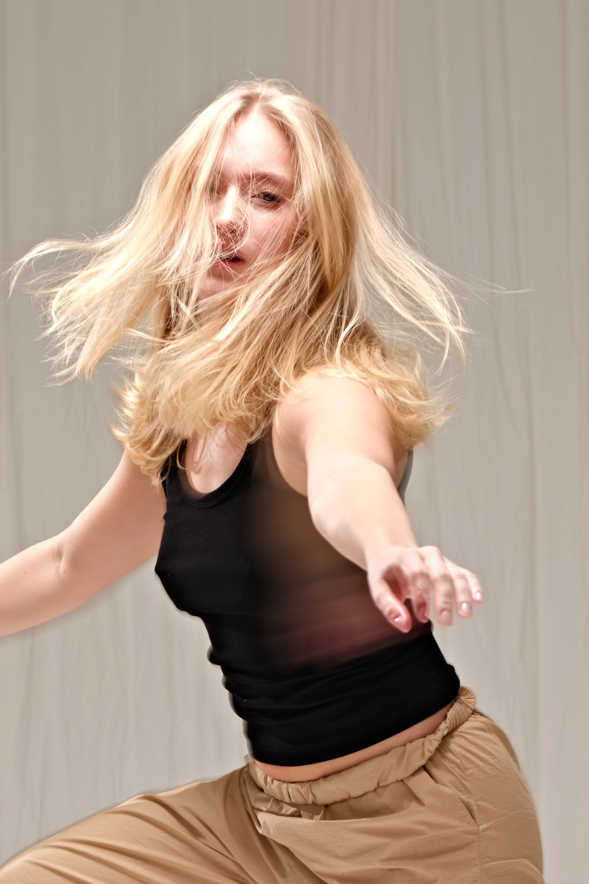 A woman with blond hair wearing a black tank top and beige pants dancing energetically with her hair flowing, against a neutral background.