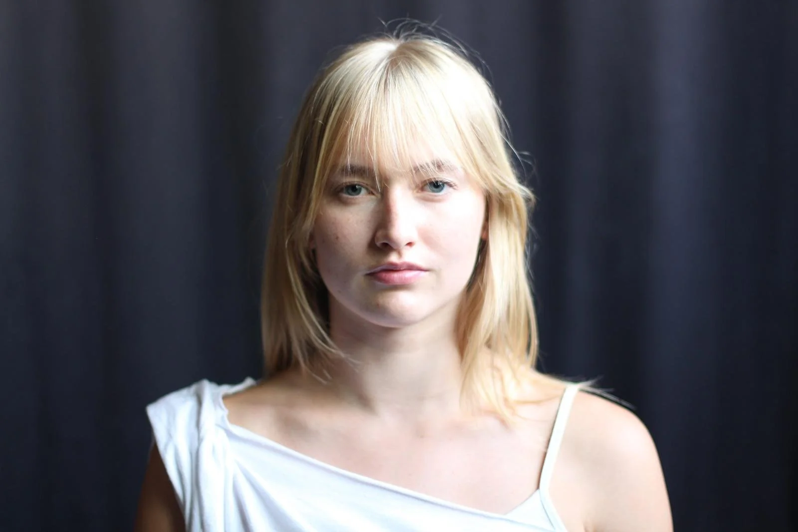 Portrait of a young woman with blonde hair and light skin, wearing a white sleeveless top, standing against a dark background.