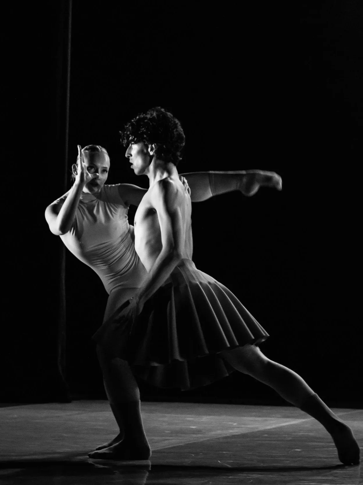 Black and white photo of two ballet dancers performing on stage. One dancer is a female with a high ponytail and a leotard, and the other is a male in a pleated skirt, with curly hair, in a dance pose.