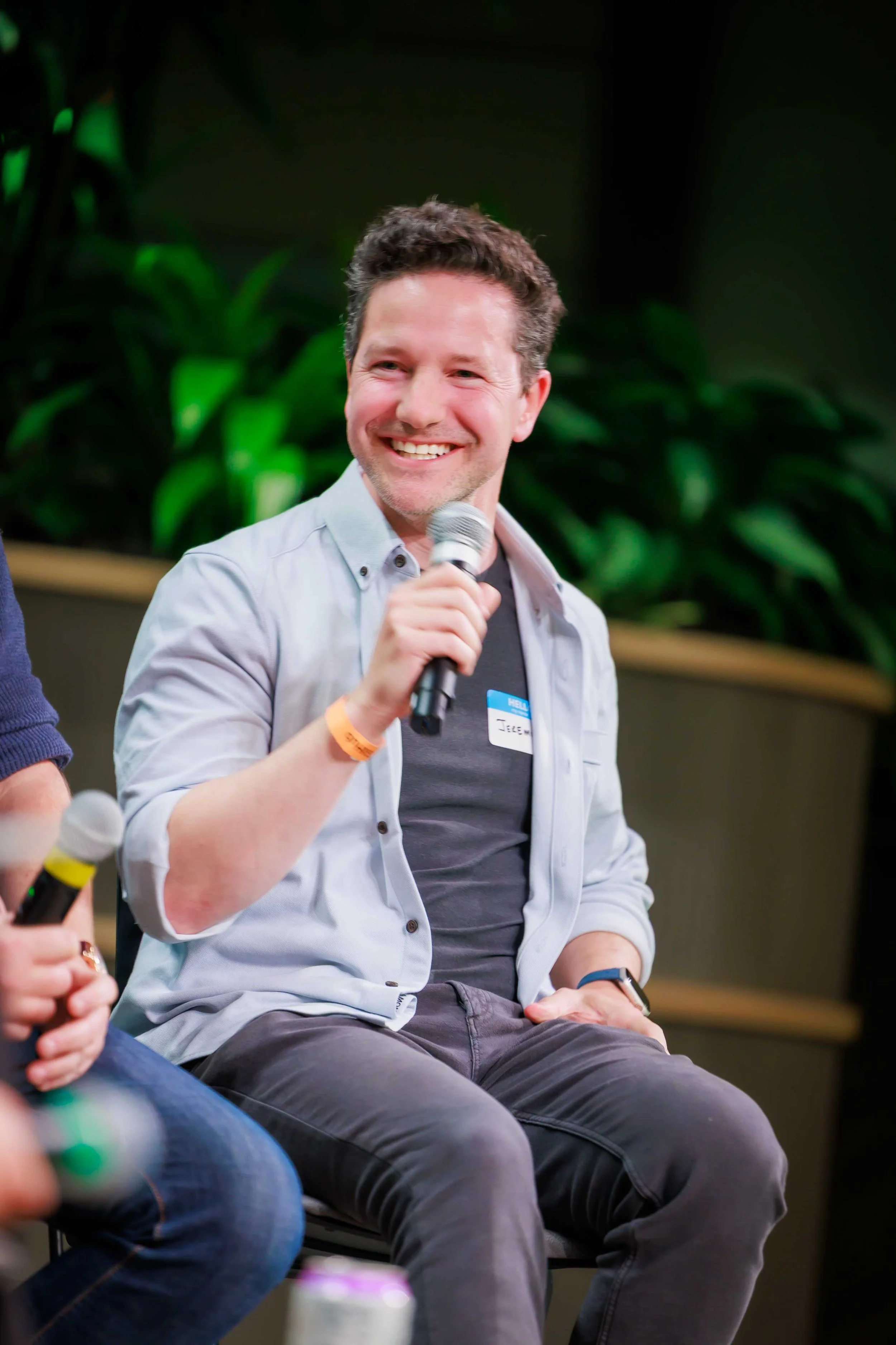 A man with dark hair, wearing a gray shirt and a light-colored jacket, smiling while holding a microphone. He appears to be participating in a panel or discussion in an indoor setting with a dark background and green plants.