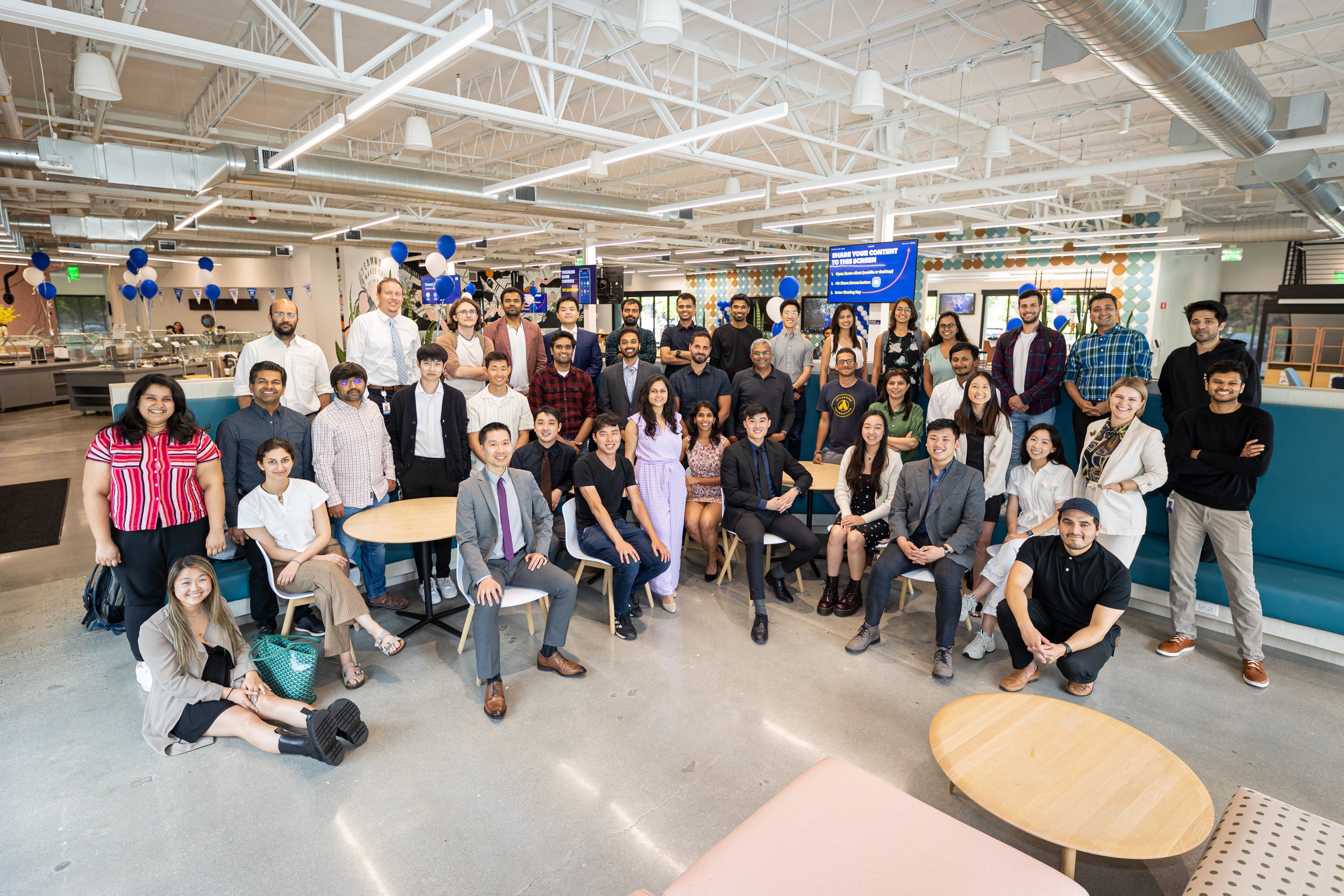 Group of diverse people gathered indoors, smiling and posing for a photo, with balloons and a digital screen in the background.