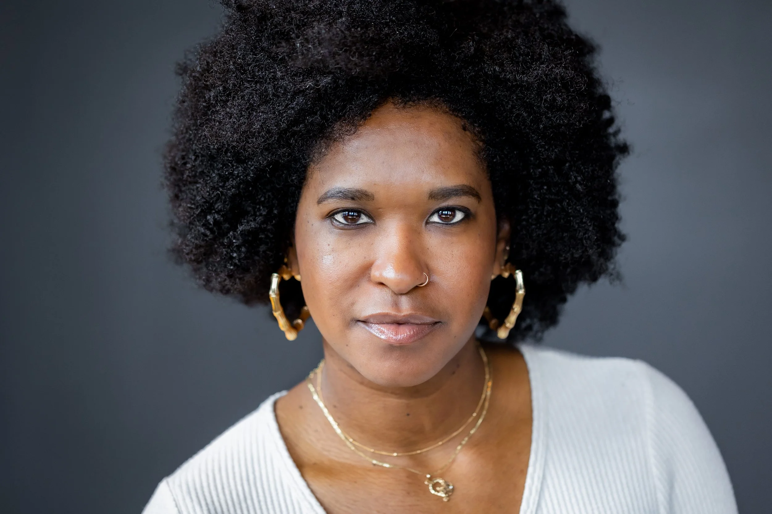Close-up portrait of an African-American woman with natural curly hair, wearing gold jewelry, a white top, and a silver necklace, against a dark background.