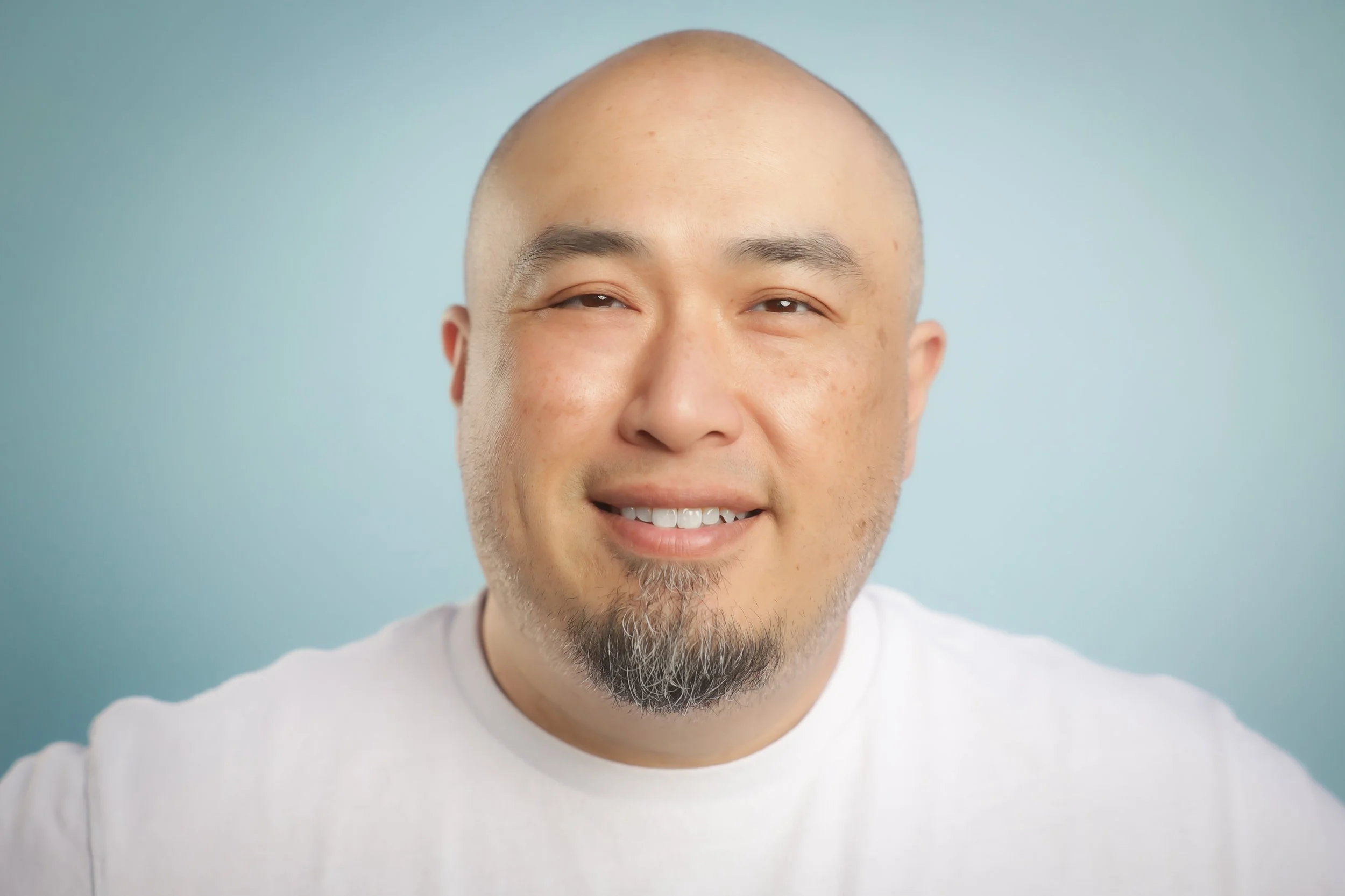 A smiling man with a bald head and a goatee, wearing a white shirt, against a light blue background.