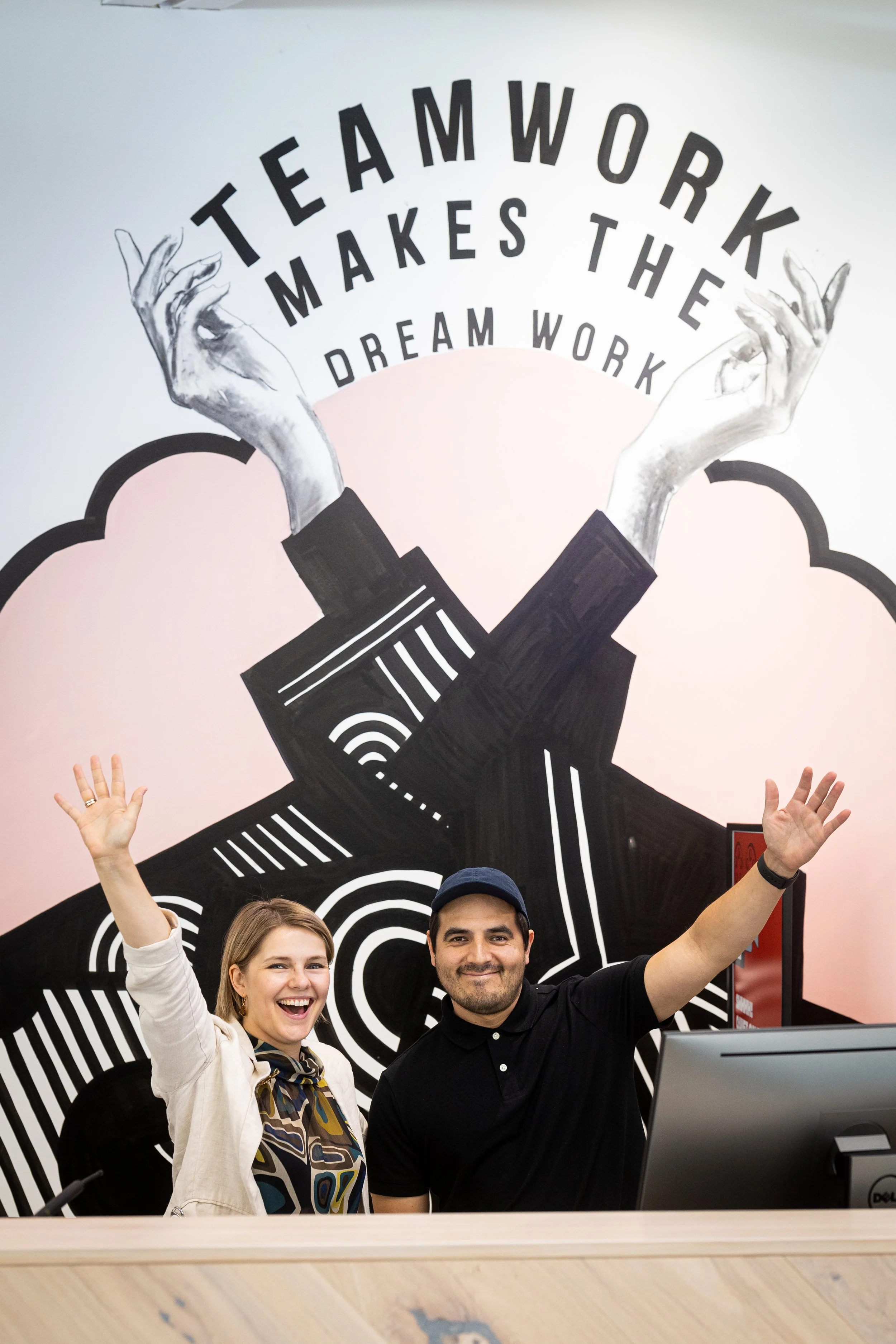 Two smiling people, a woman and a man, are cheering with their hands raised in front of a mural with the words 'Teamwork Makes the Dream Work' and a pair of stylized hands.