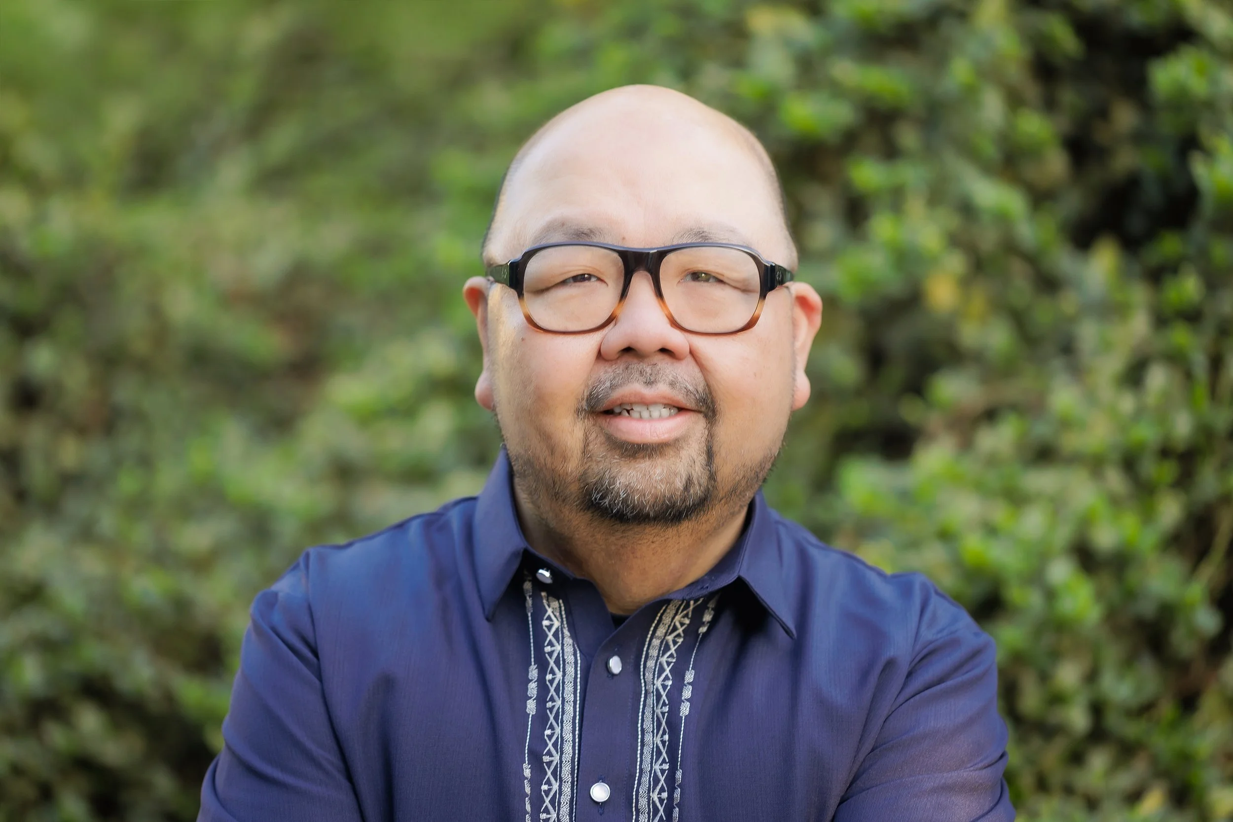 A bald man with glasses smiling in front of green foliage, wearing a navy blue shirt with embroidered patterns.