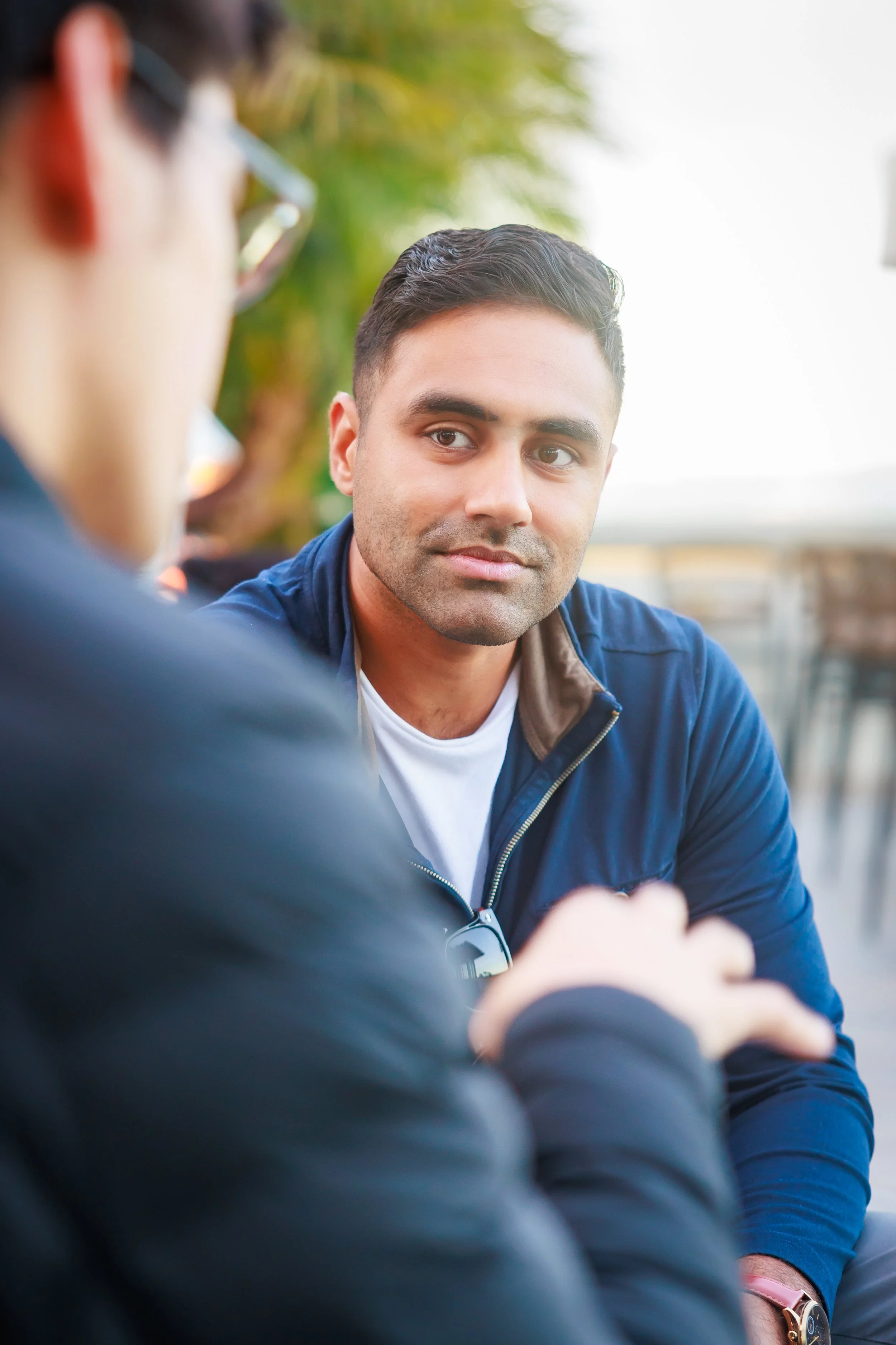 A young man with short, dark hair and a light complexion is sitting outdoors and looking at another person who is partially visible and out of focus in the foreground. The background features green foliage and a bright sky.