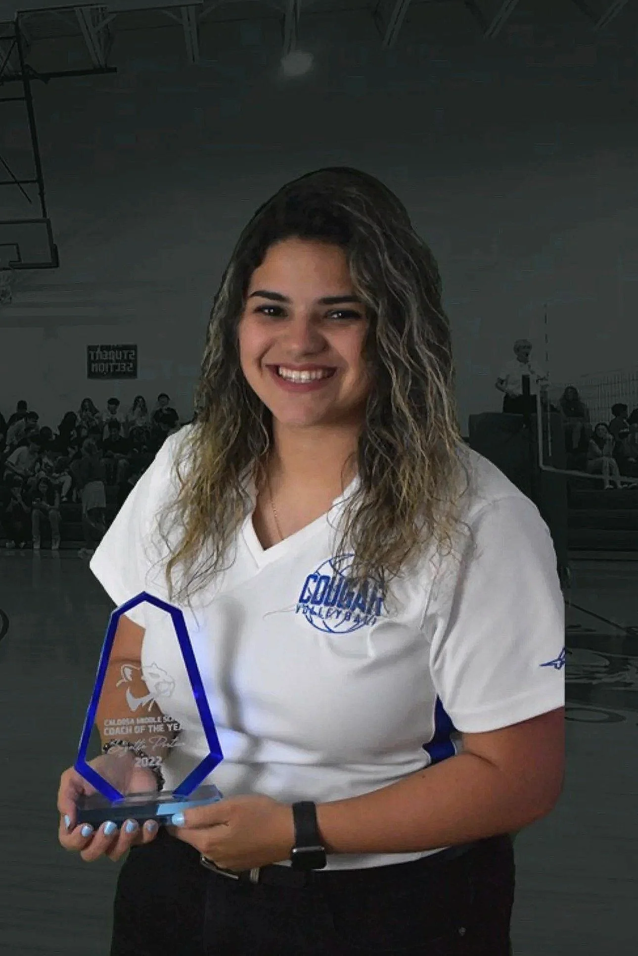A young woman with curly hair smiling while holding a glass award in a gymnasium with other people sitting on bleachers in the background.