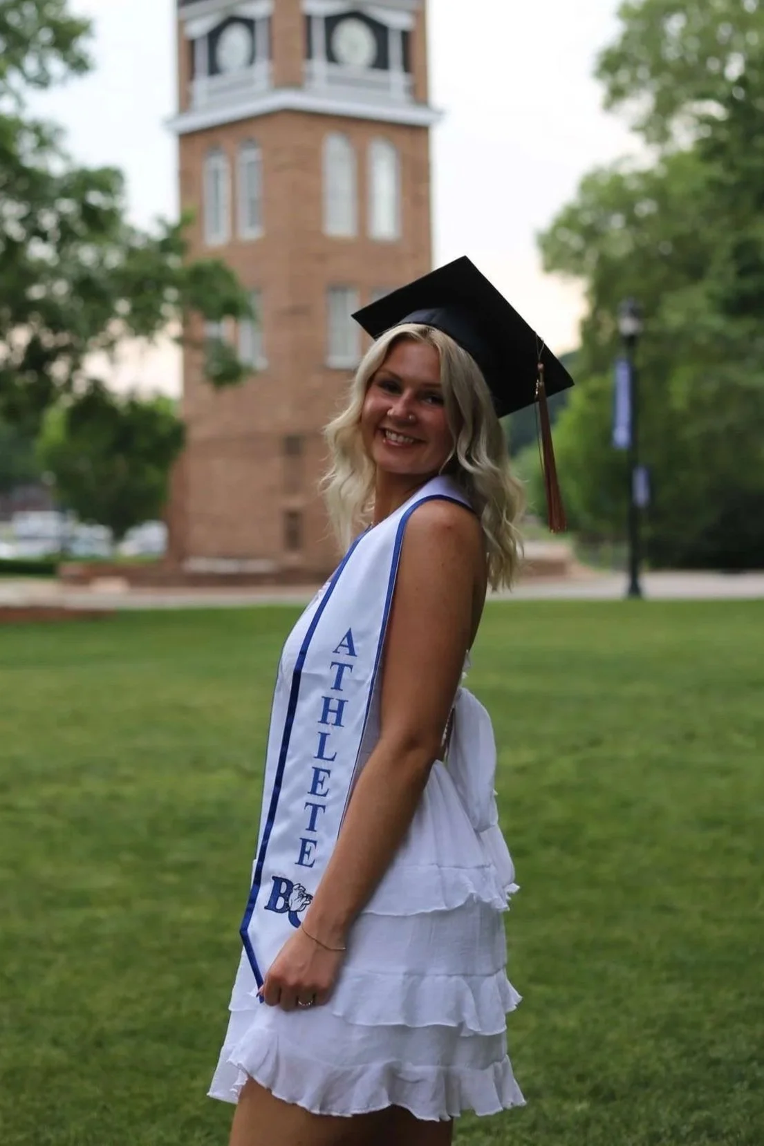A young woman wearing a graduation cap and white dress, smiling outdoors with a brick tower and green trees in the background.