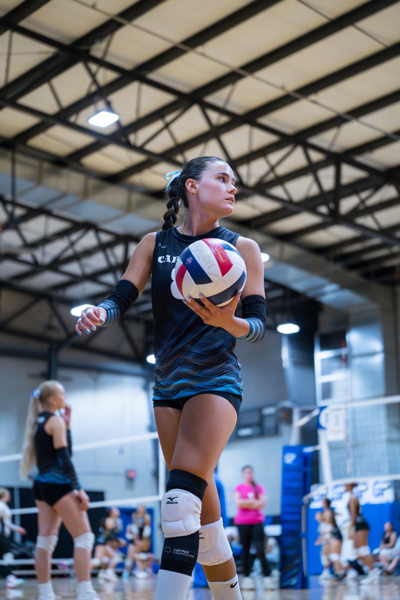 A female volleyball player holding a volleyball during practice or a game in an indoor gymnasium. She is wearing a black sleeveless jersey, knee pads, and black shorts, with her hair in a braid and focused on the game.