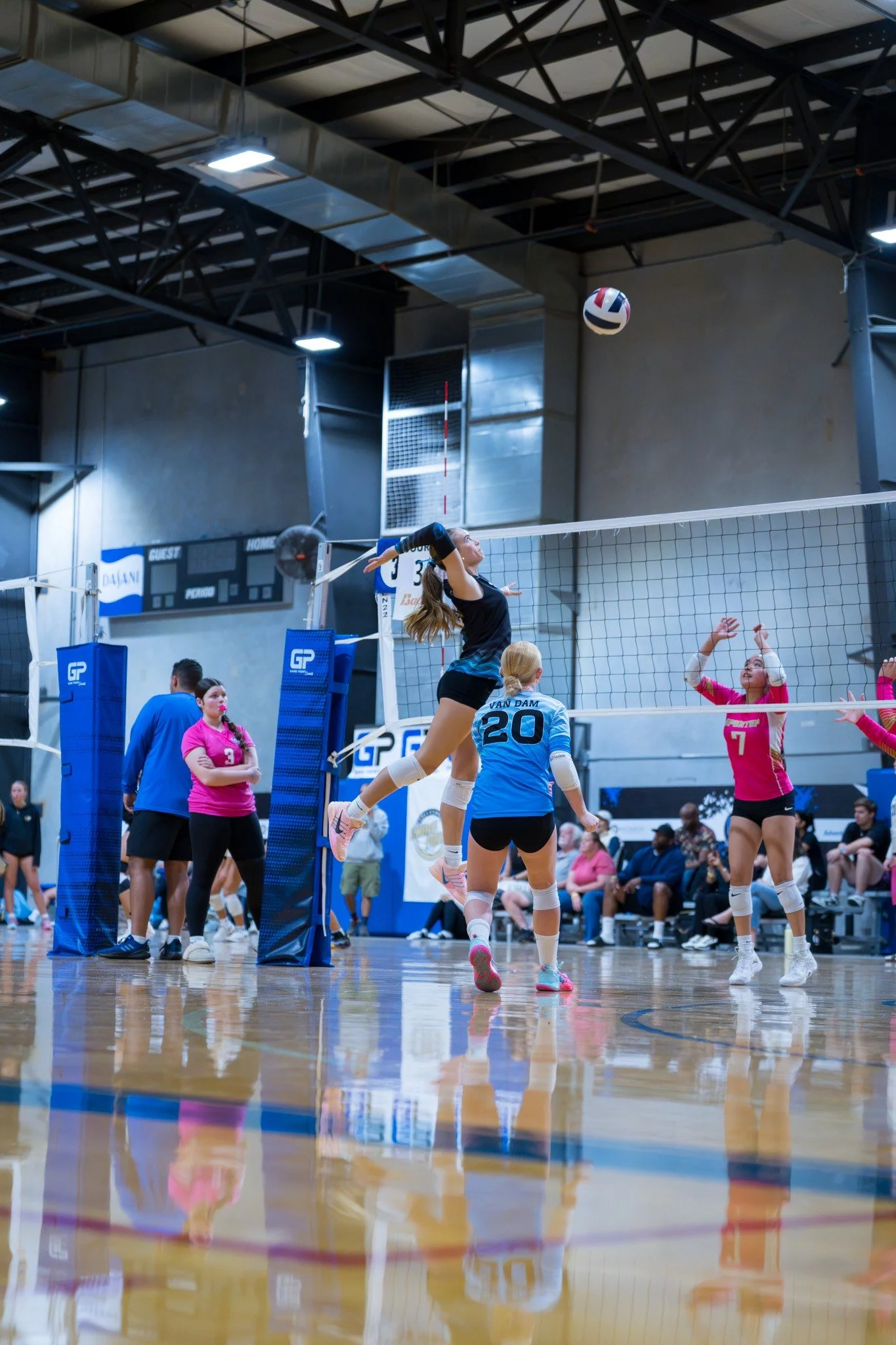 A volleyball game in progress with a player jumping to hit the ball over the net, while others on the court and audience watch.