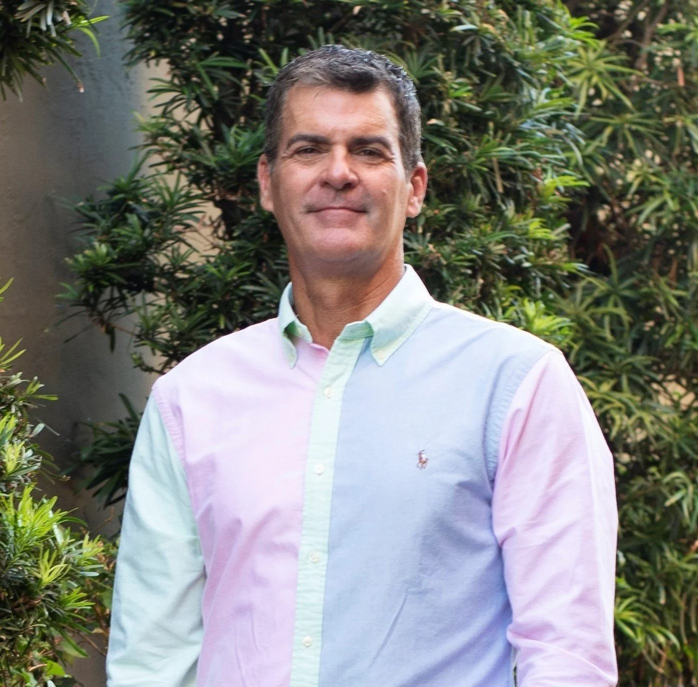 A man with short dark hair wearing a pastel-colored button-up shirt standing outdoors with greenery in the background.