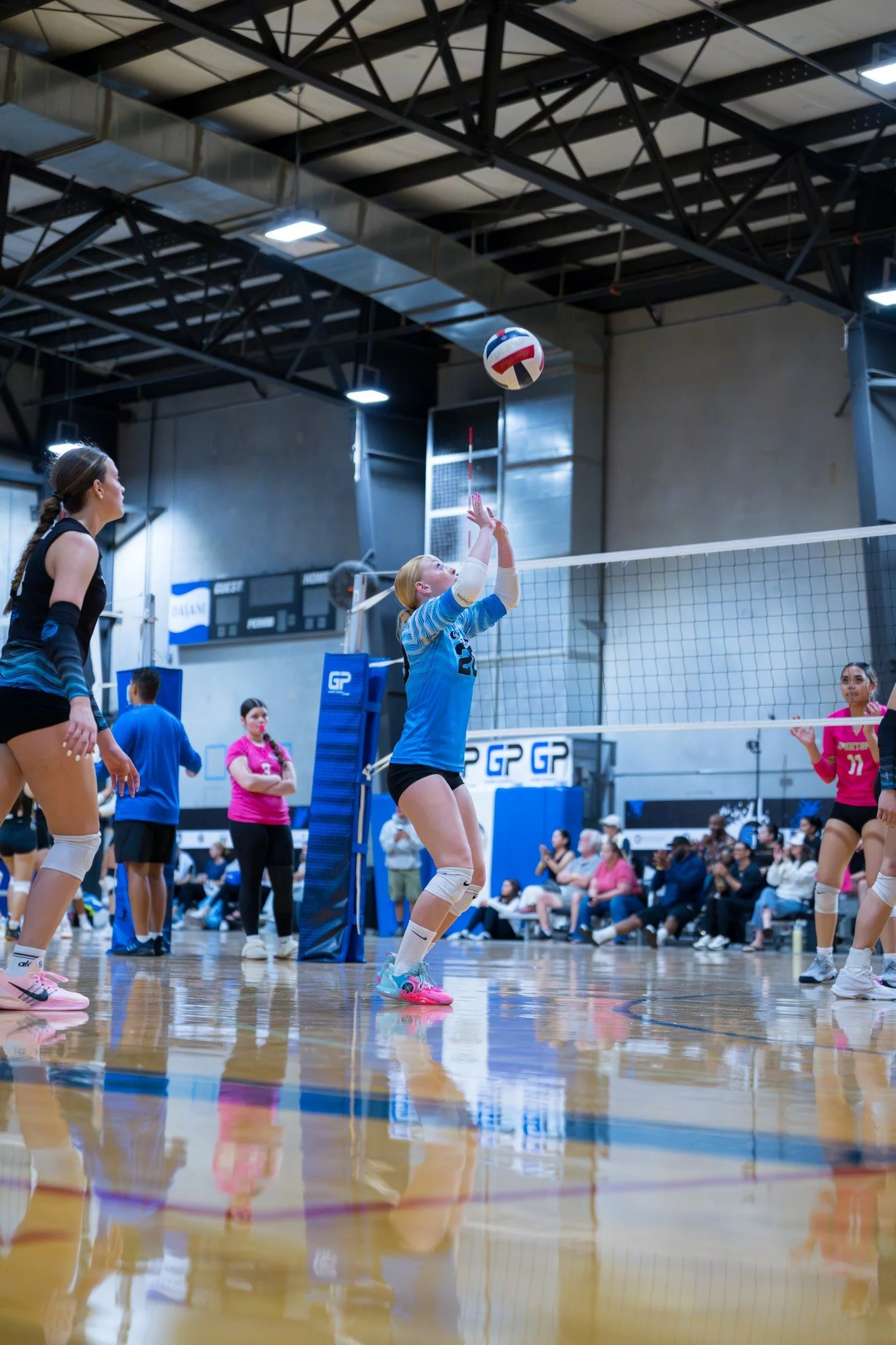 A volleyball game in progress inside an indoor gym, with players preparing to hit or serve the ball over the net, surrounded by spectators.