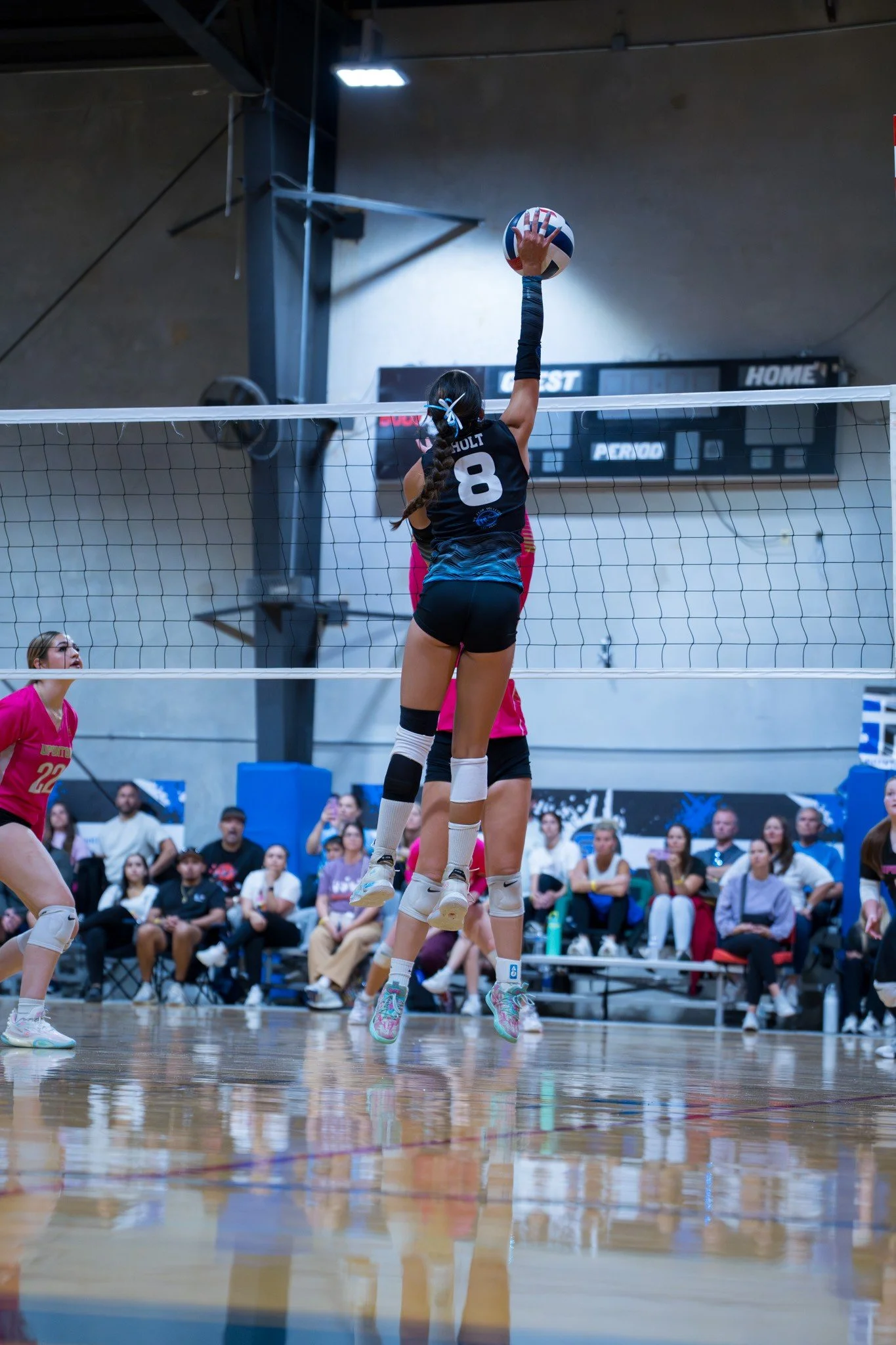 A female volleyball player wearing a black uniform with the number 8 is jumping to hit the ball over the net during a game, with spectators watching from the bleachers in the background.