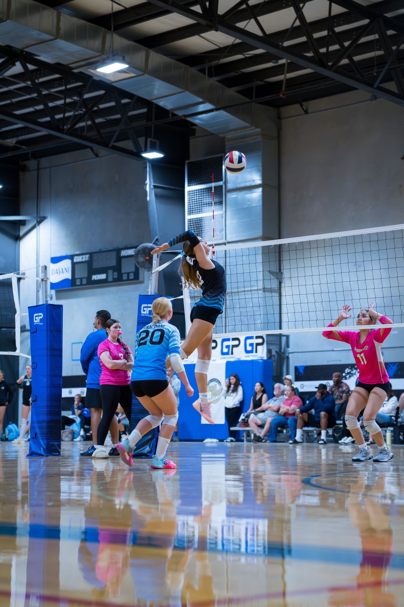 Indoor volleyball game with players at the net, one player in a black uniform jumping to hit the ball, audience seated in the background, and scoreboard visible.