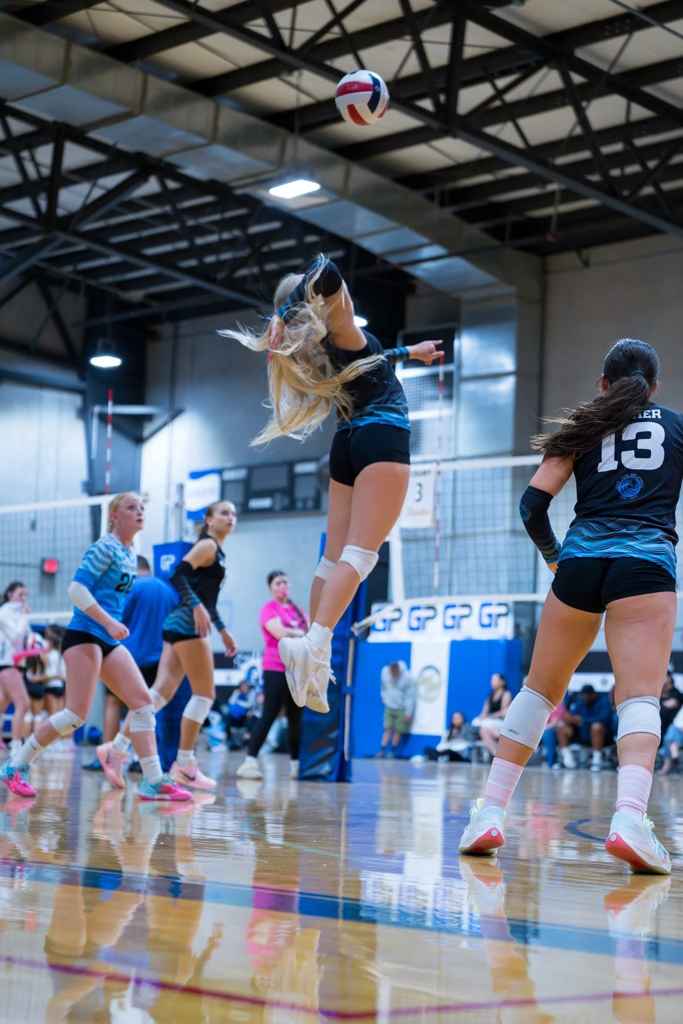 A women's volleyball game is in progress inside a gymnasium with a high ceiling. One player wearing a black and blue uniform is mid-air preparing to spike the ball. Other players on the court are watching or getting ready to react.