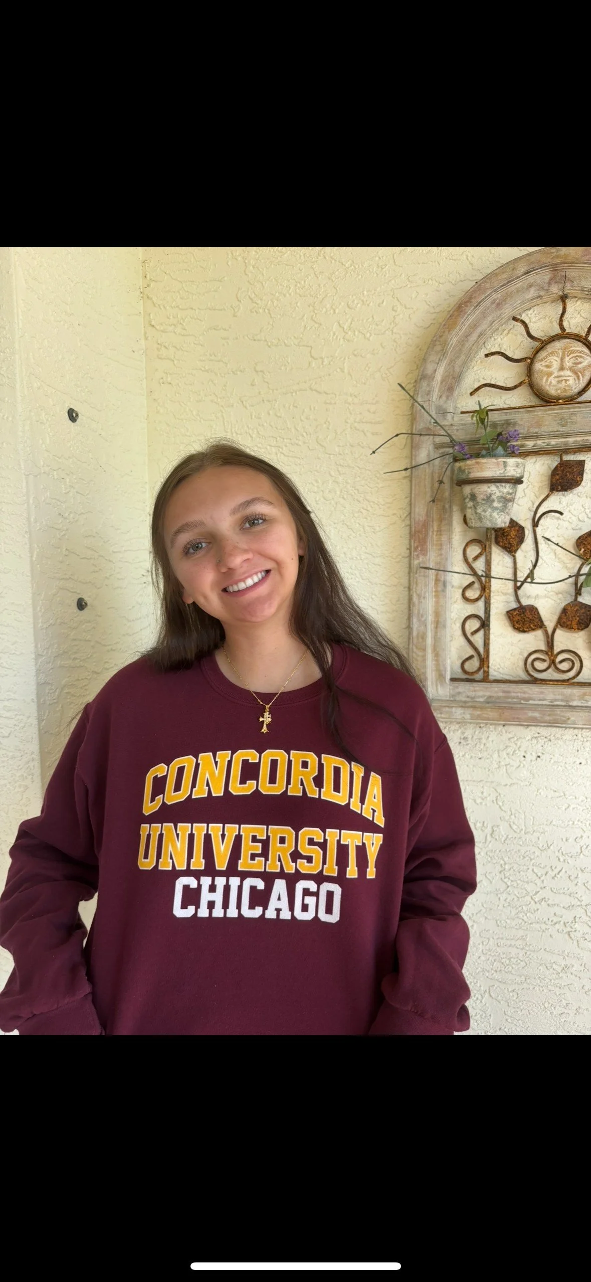A smiling young woman with long brown hair wearing a maroon sweatshirt with yellow and white text that reads "Concordia University Chicago". She is standing in front of a textured light yellow wall with a decorative metal sun and plant wall hanging.