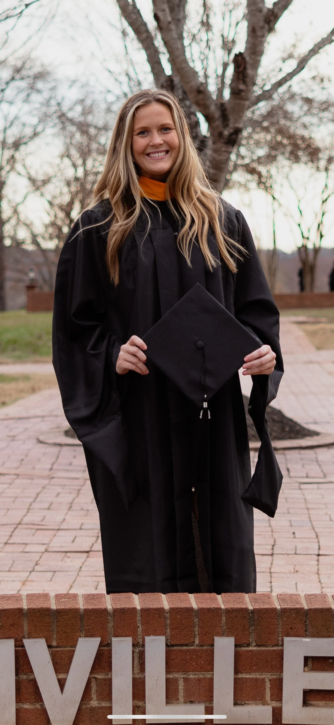 A young woman in a black graduation gown holding a mortarboard, standing outdoors on a brick walkway with trees in the background, smiling.